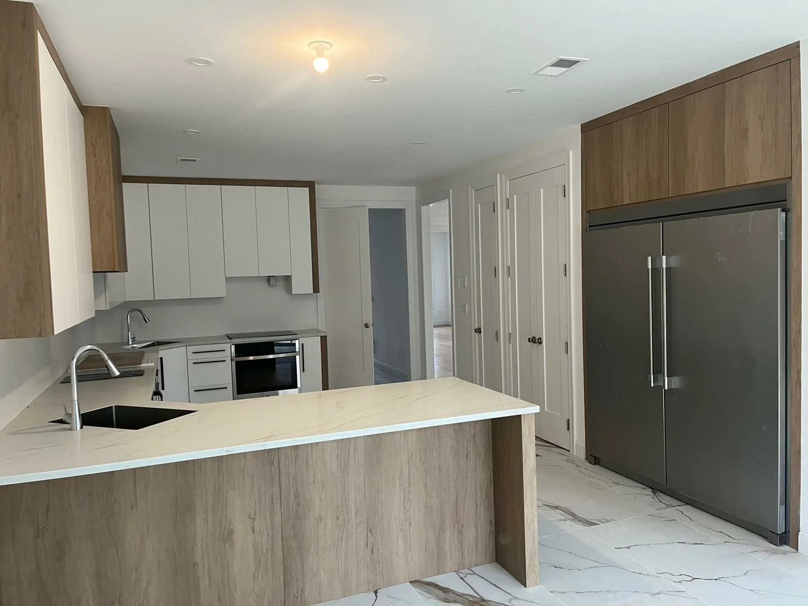 Modern kitchen with white and wood cabinets, stainless steel refrigerator, marble countertop, and tile floor.