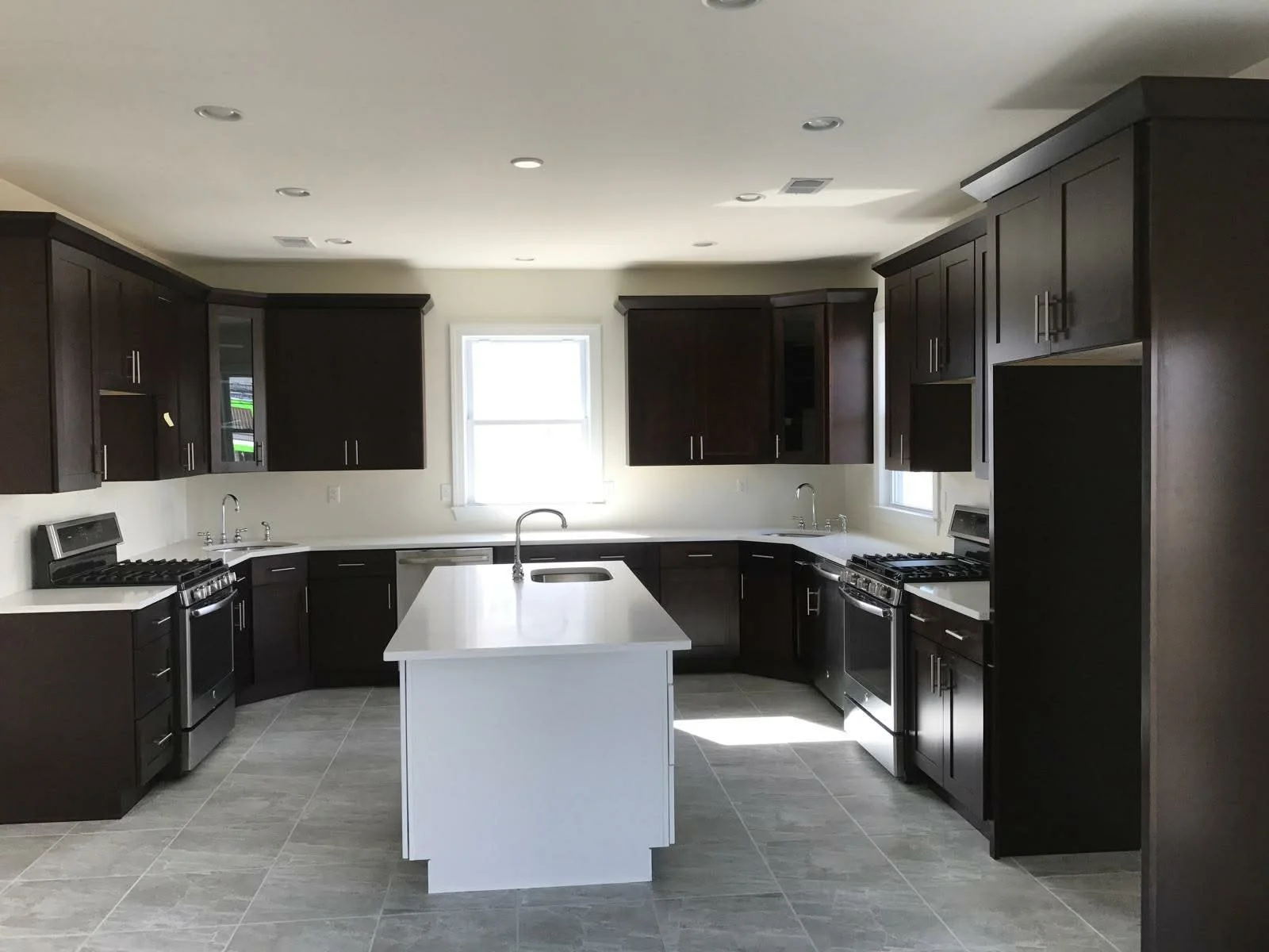 Modern kitchen with dark wood cabinets, white countertops, a white island in the center, stainless steel appliances, and two windows providing natural light.