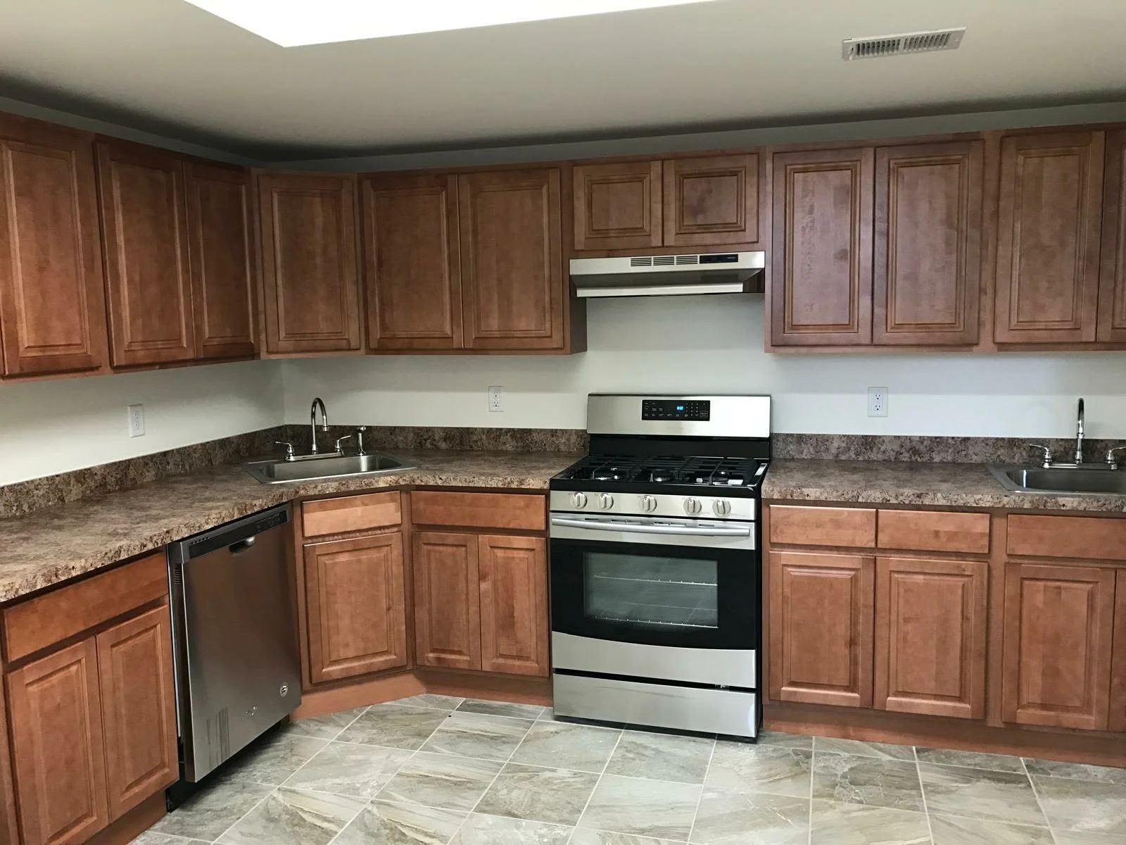 Kitchen with brown cabinets, stainless steel dishwasher, black and stainless steel stove, granite countertops, double sinks, and tiled floor.