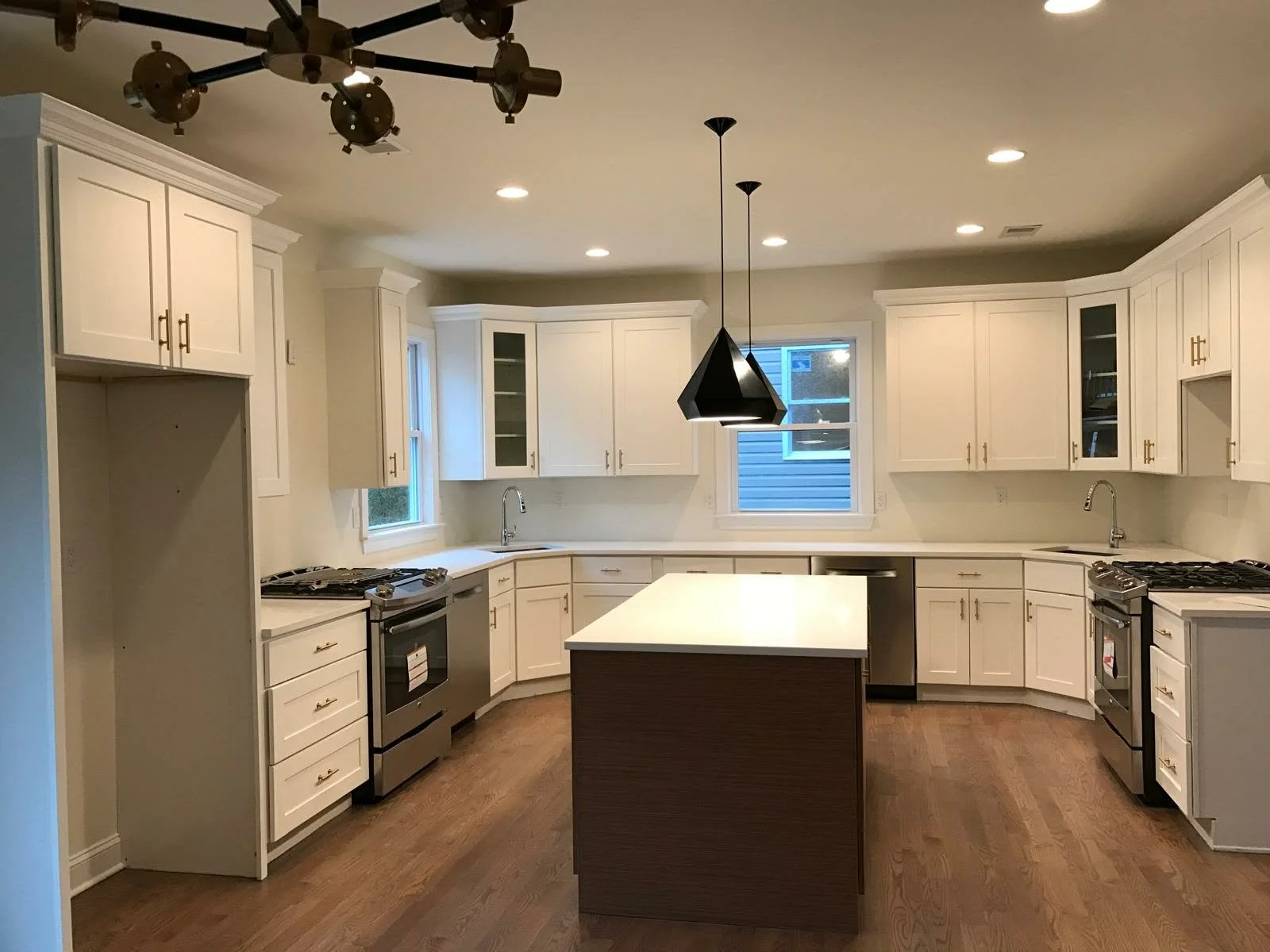 A modern kitchen with white cabinets, hardwood floor, and a central island with a dark wood base. It has three windows, two gas stoves, and a black pendant light hanging over the island.