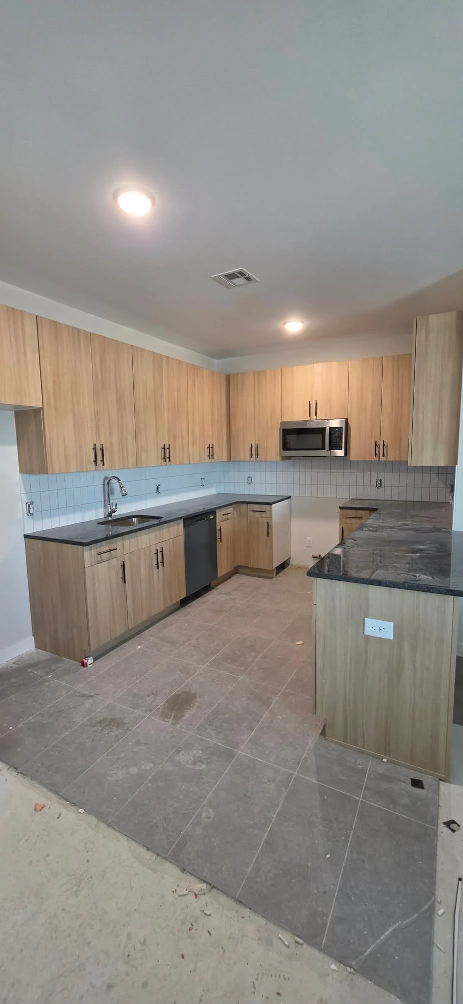 Modern kitchen with wooden cabinets, black countertops, stainless steel appliances including a microwave and dishwasher, white tile backsplash, and unfinished flooring.