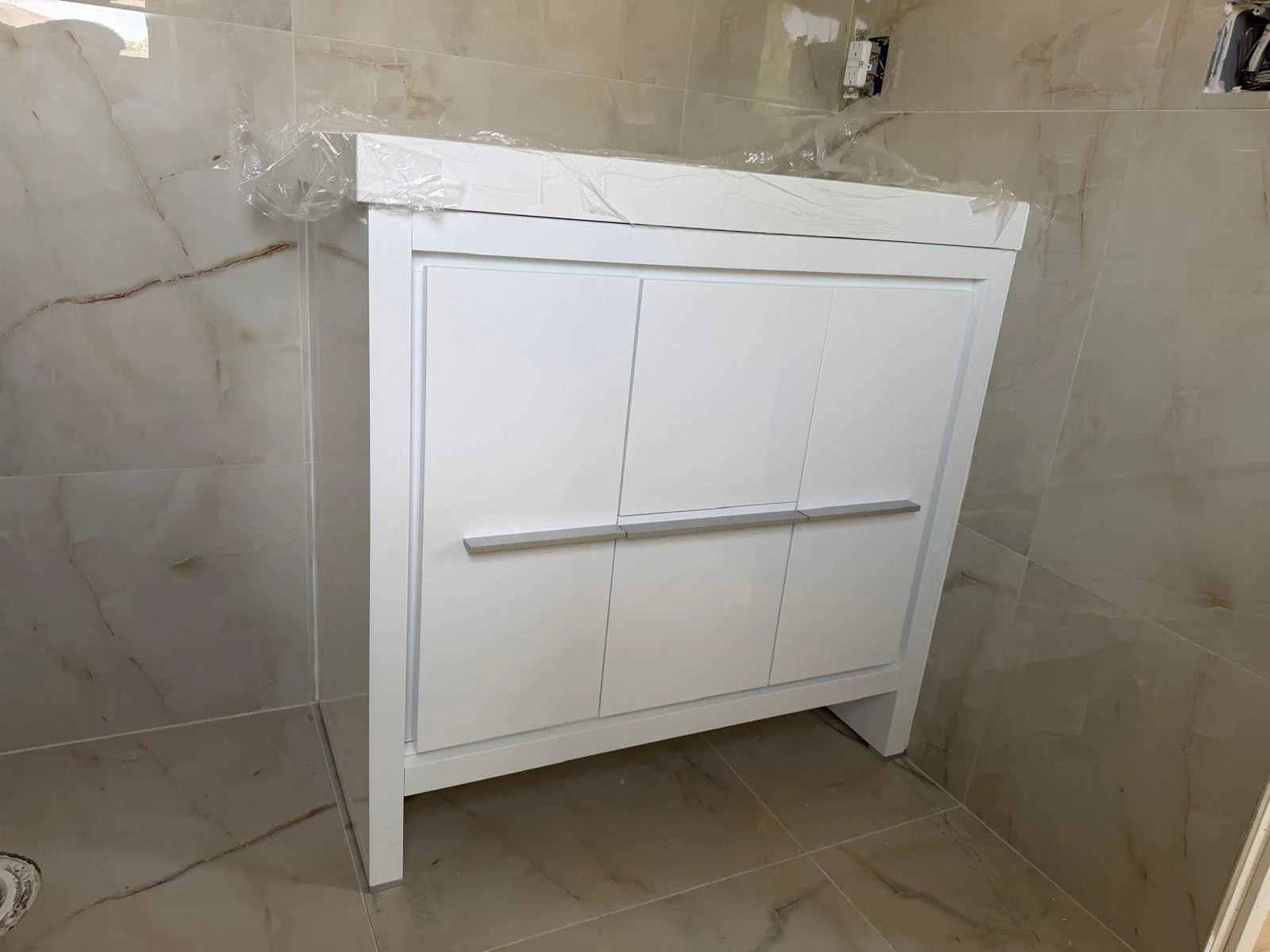 White bathroom vanity cabinet with a metal handle, placed against a marble wall and floor.