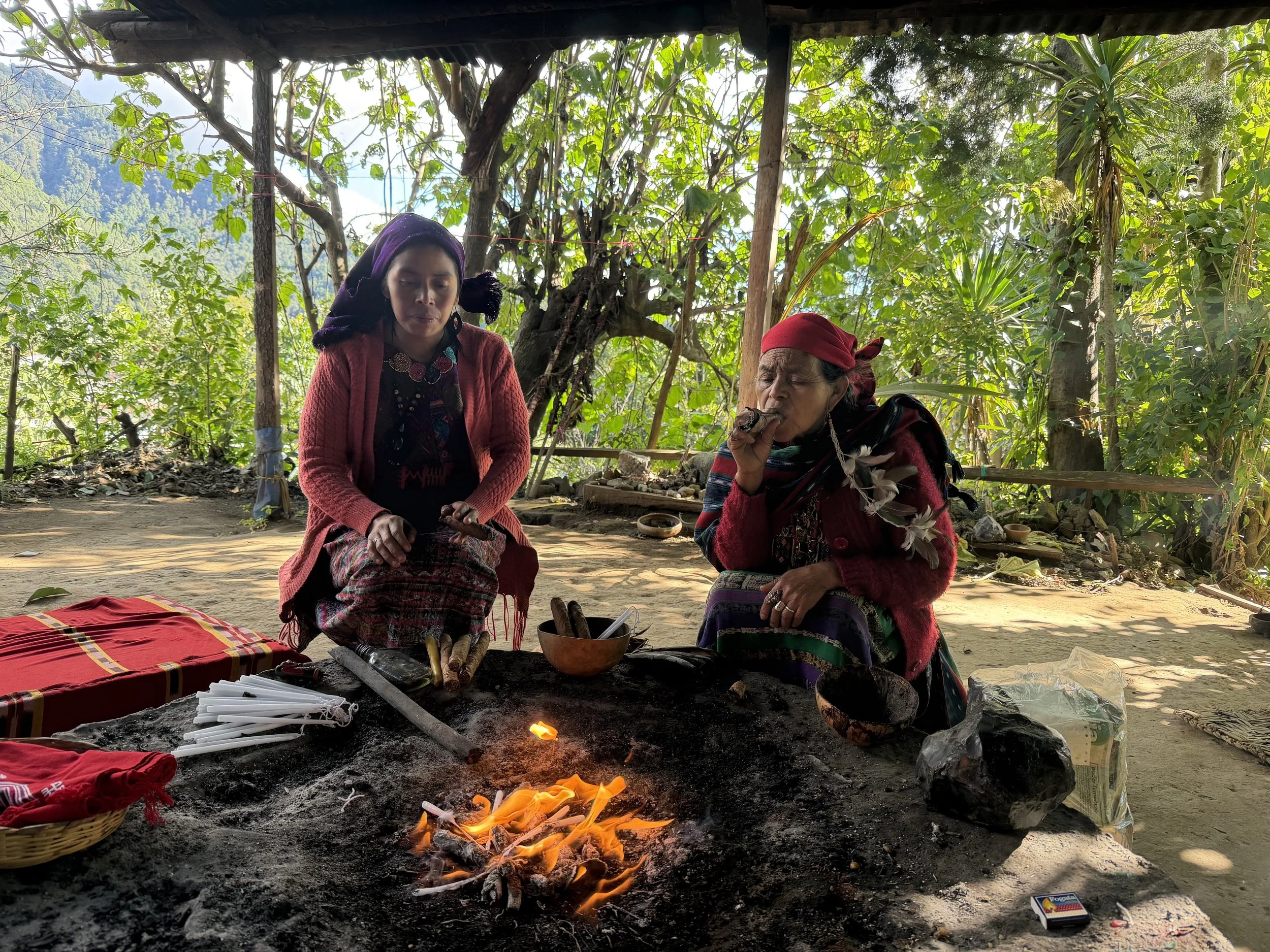 Two women dressed in traditional clothing sitting around an open fire, with one of them smoking and the other looking down, surrounded by lush green trees.