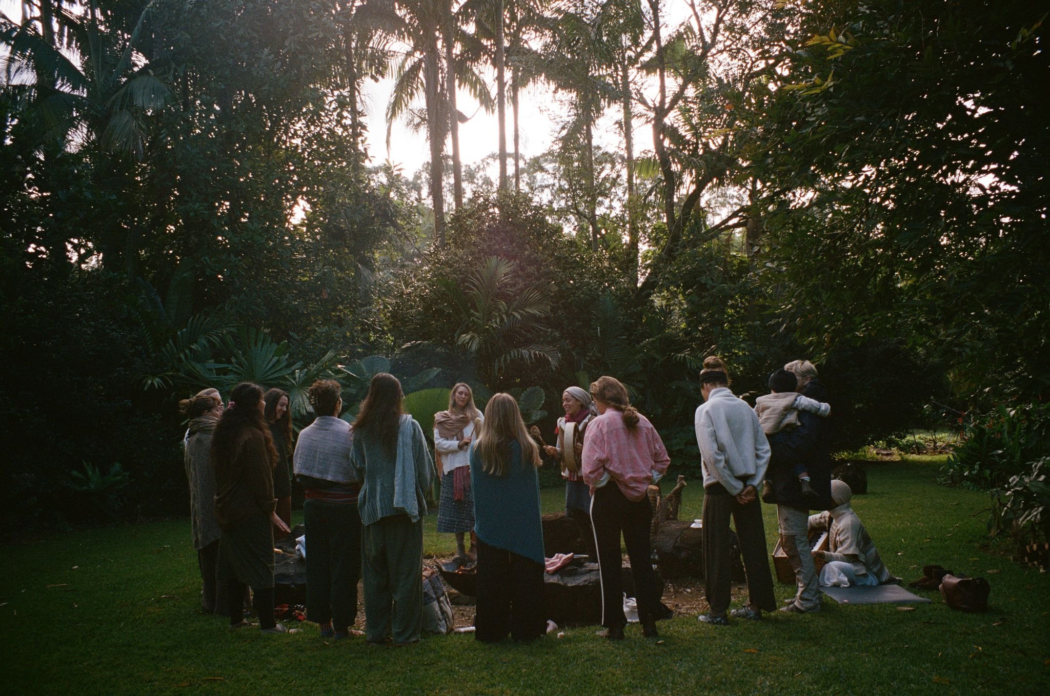 Group of people participating in a meditation or yoga session outdoors in a lush, green environment with tall trees and sunlight filtering through the foliage.