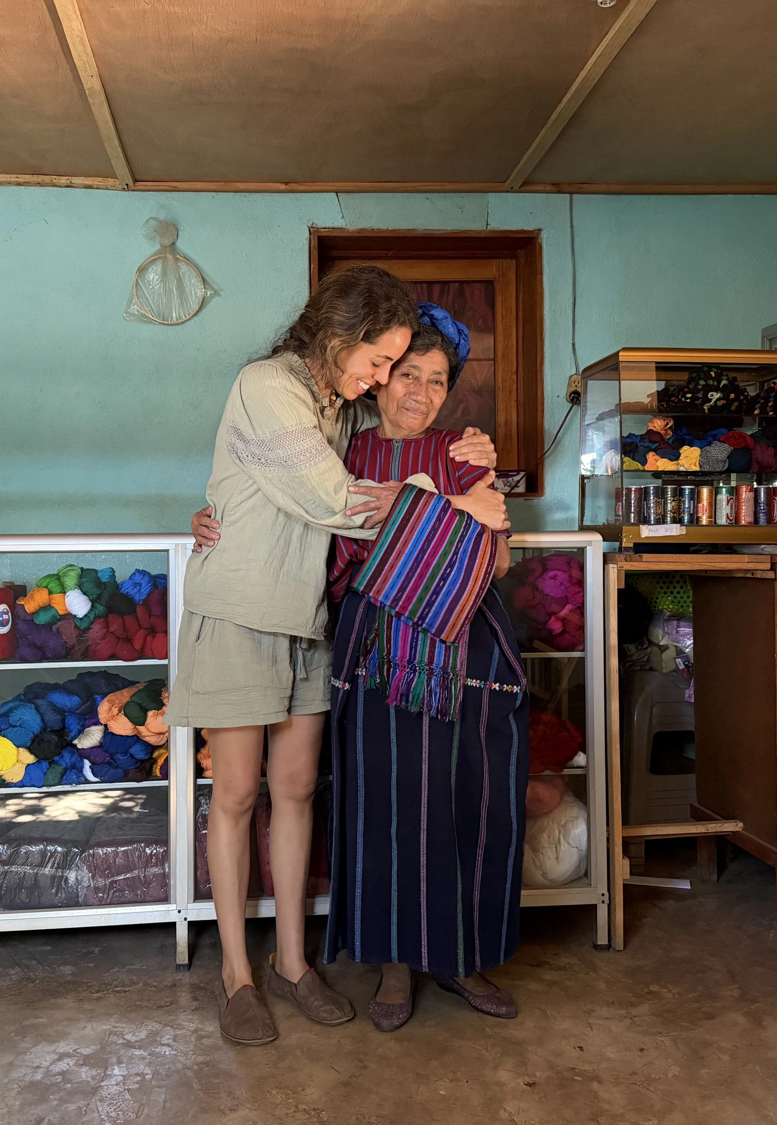 Two women hugging inside a room with shelves of yarn and textiles in the background. One woman is in traditional indigenous clothing and the other in casual Western clothing.