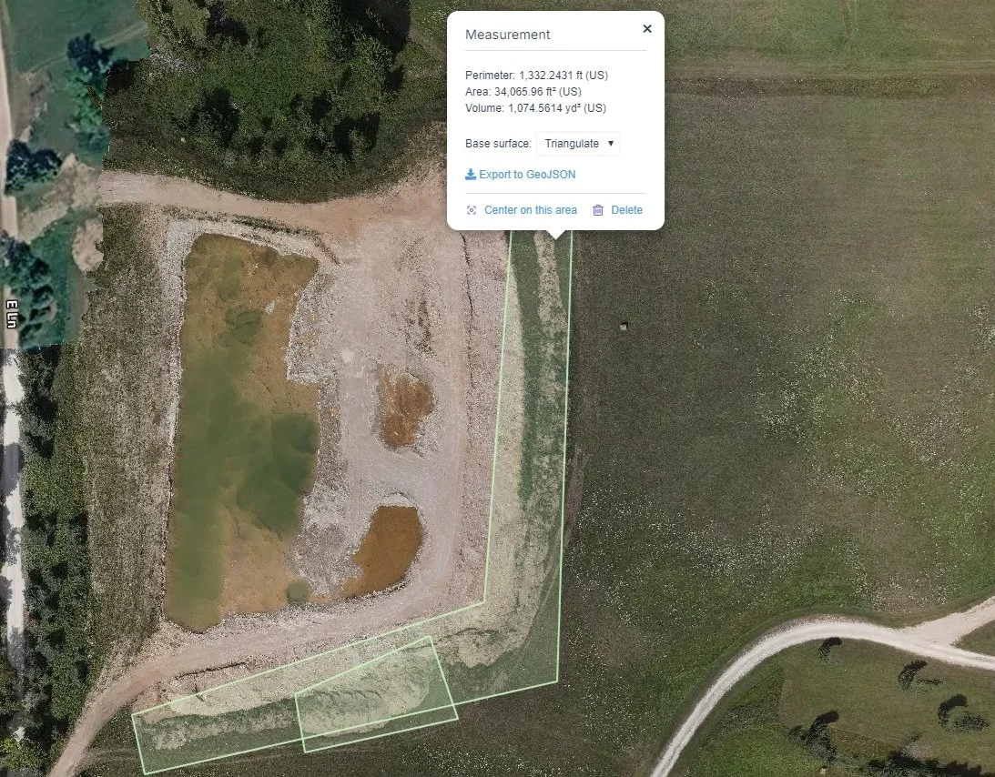 An aerial view of a construction site or cleared area with patches of grass and dirt, surrounded by trees and some developed land with roads on the sides.