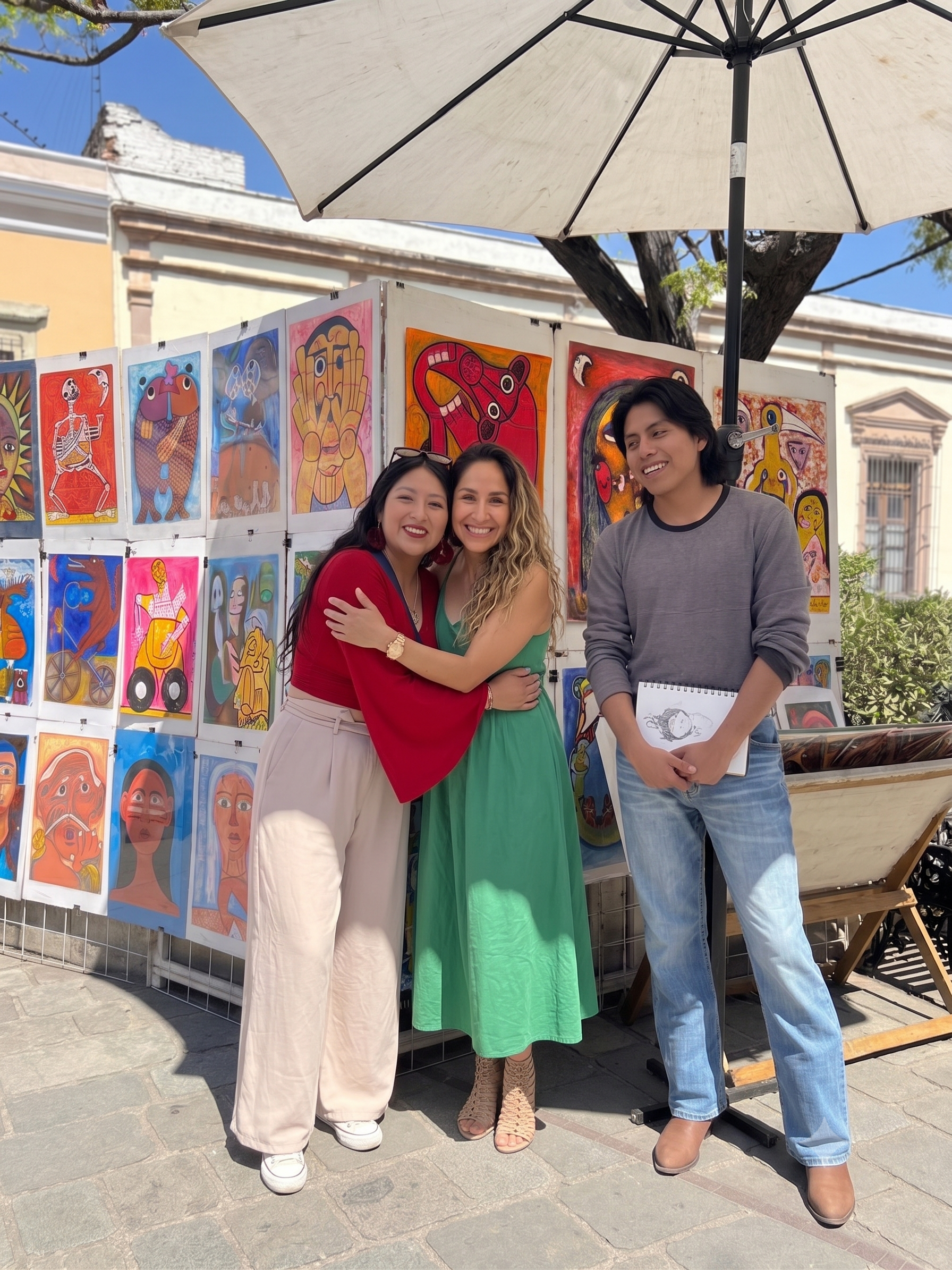 Tres personas sonriendo y abrazándose frente a un puesto de arte con pinturas coloridas en exhibición bajo una sombrilla en la calle.