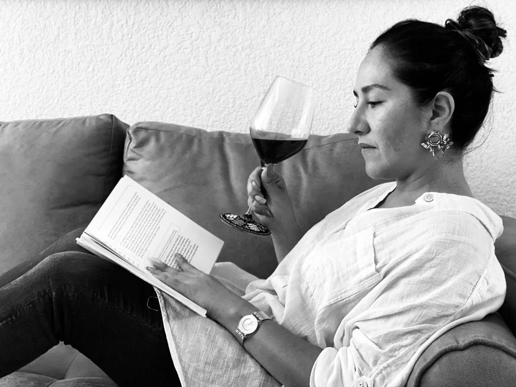 Mujer sentada en un sillón con un libro en las manos, sosteniendo una copa de vino tinto.