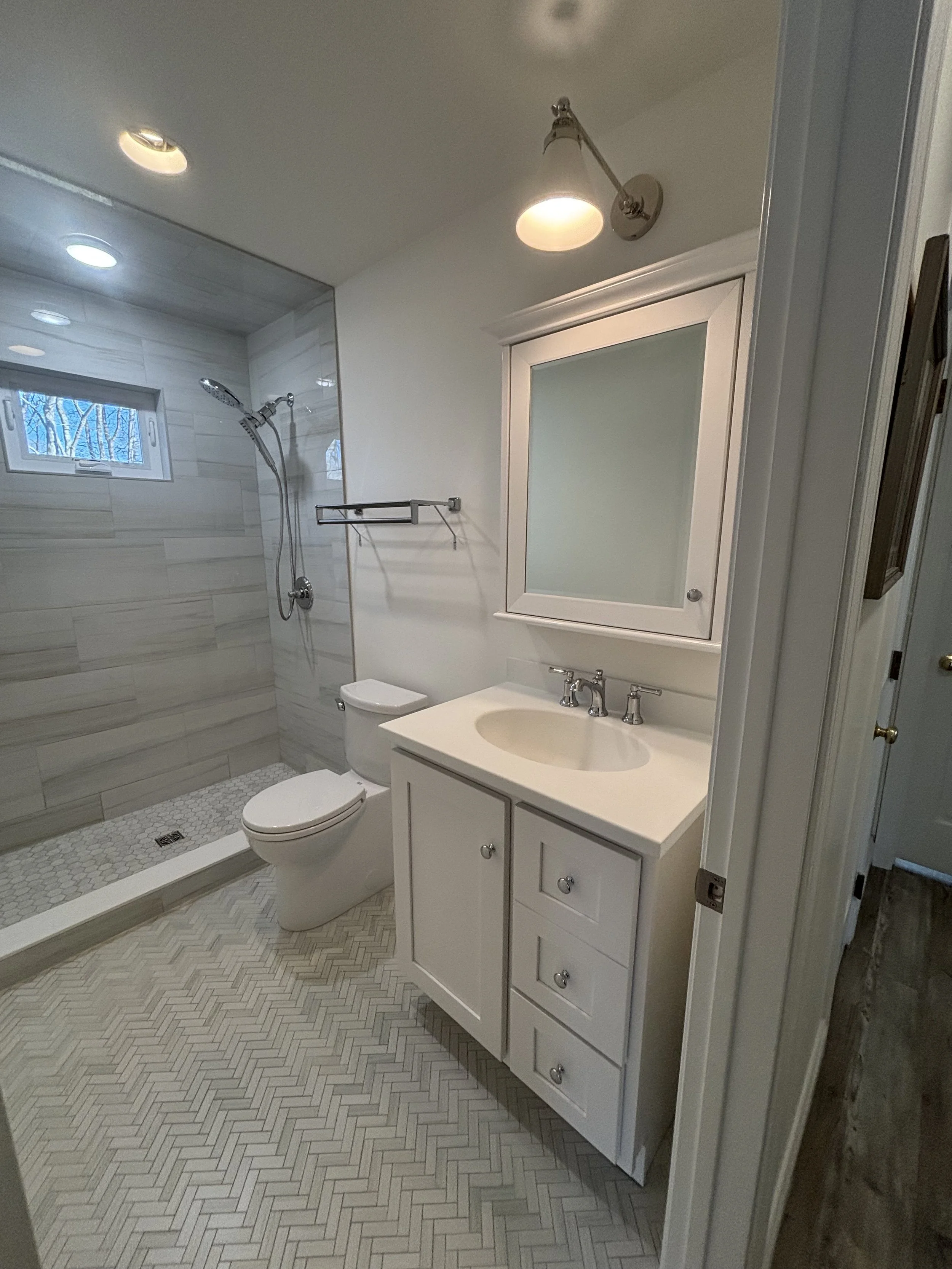 Completed bathroom renovation with tiled shower, vanity, toilet, and herringbone-pattern tile flooring.