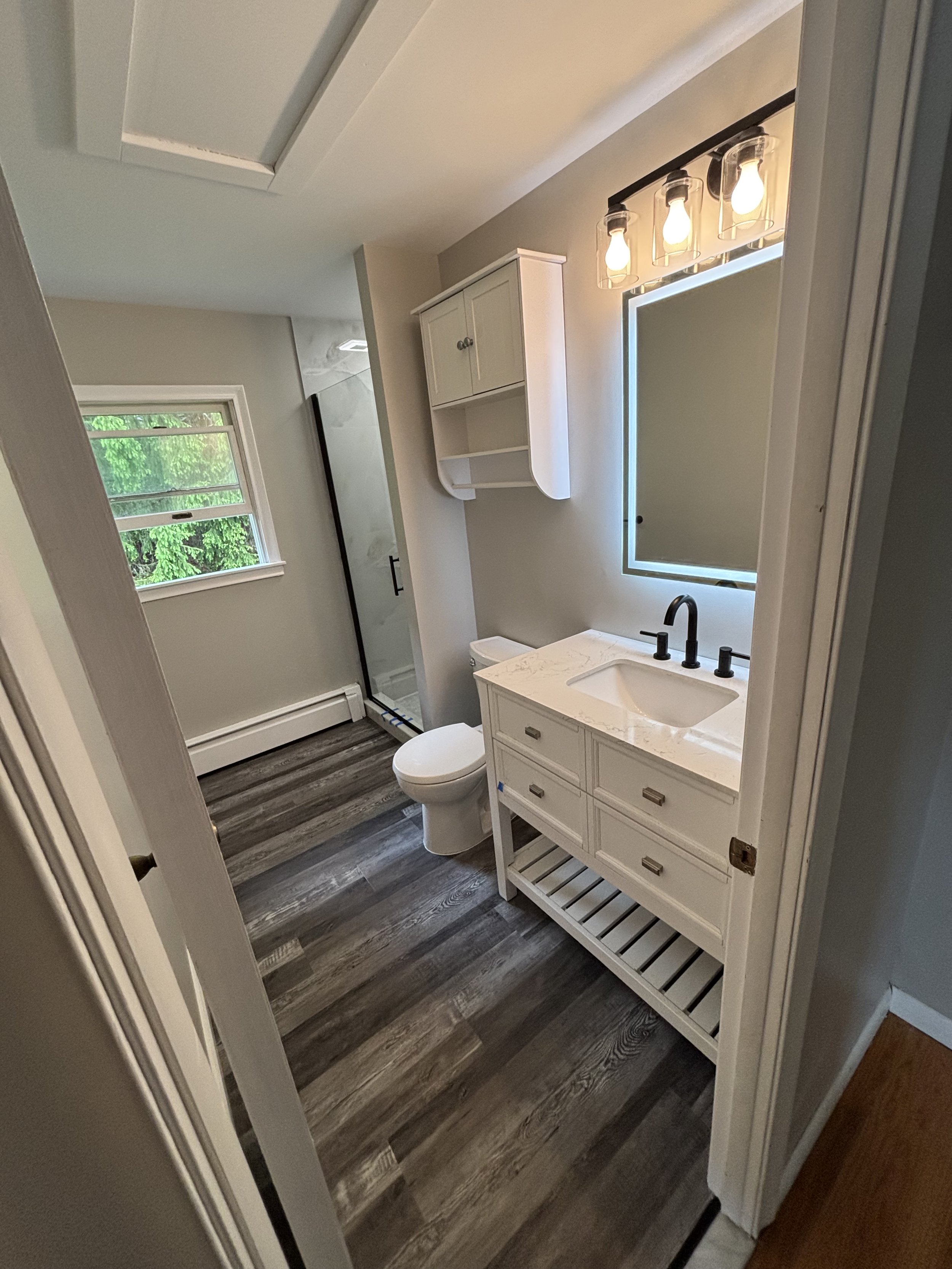 Renovated bathroom with vanity, illuminated mirror, glass enclosure, and wood-look flooring.