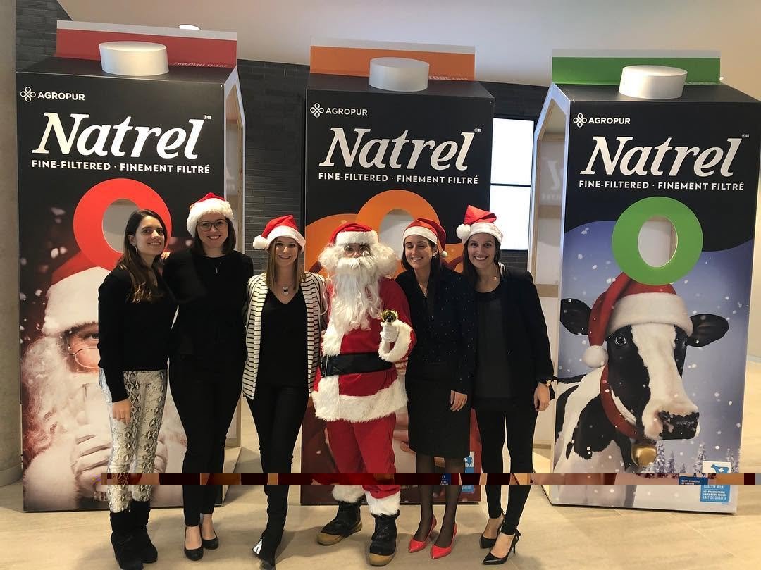 Groupe de six personnes, dont un Père Noël, posant devant des affiches de la marque Natrel avec des images de vaches et des décorations de Noël.