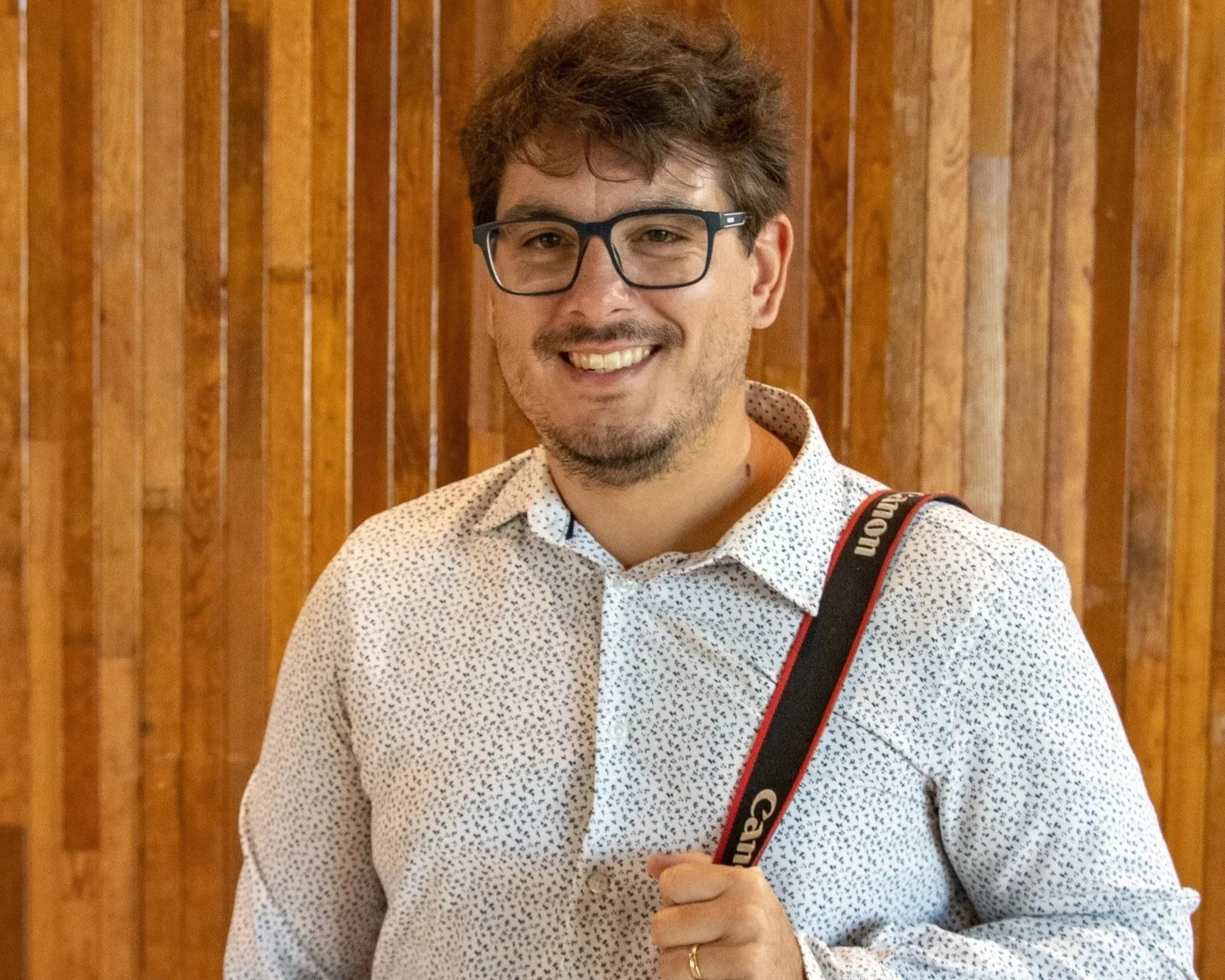 Le fondateur de l'agence, souriant, portant des lunettes et une chemise à motifs, avec sa caméra en bandoulière sur l'épaule, se tient devant un mur en bois.