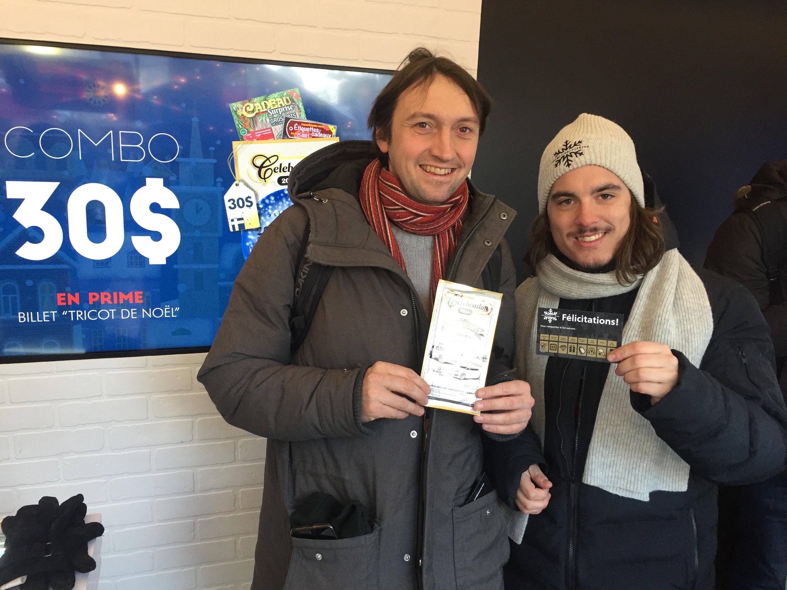 Deux hommes souriants posent pour une photo, l'un tient un billet de loterie ou de jeu, ils sont vêtus chaudement avec des bonnets et écharpes, à côté d'un mur blanc avec une affiche promotionnelle en arrière-plan.