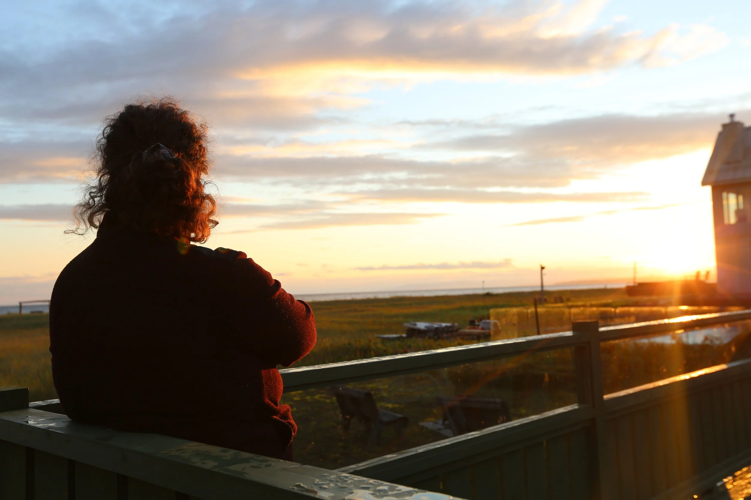 Une personne accotée sur une clôture regarde le coucher de soleil sur la mer, avec un ciel coloré et quelques nuages, près d'une maison en bois à droite.