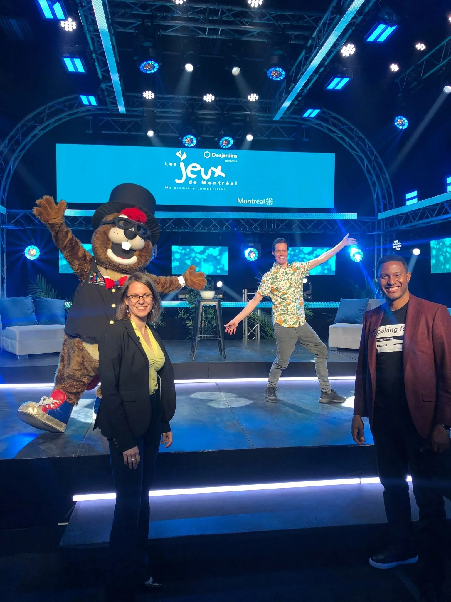 Un groupe de personnes avec une mascotte, lors d'un événement présenté par Les Jeux de Montréal. La scène est éclairée avec des lumières bleues et il y a un grand écran derrière avec le logo de l'événement.