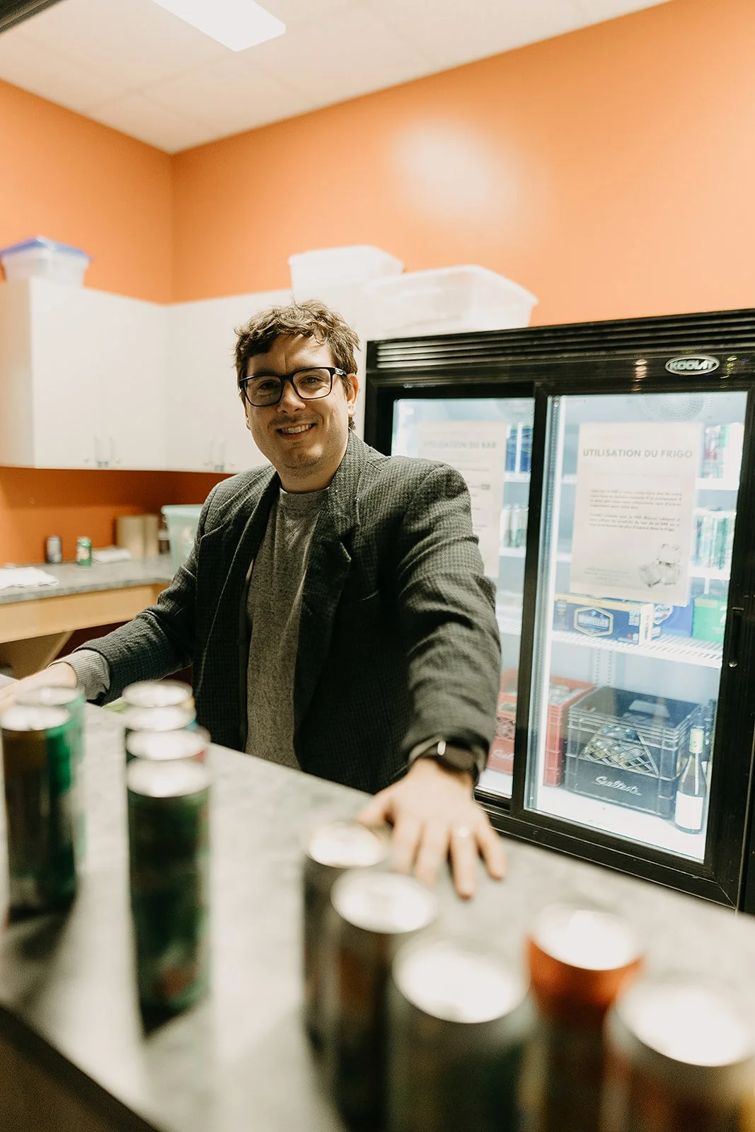 Le fondateur de l'agence, souriant derrière un comptoir de bar, avec des canettes en avant, dans un environnement intérieur, lors d'une conférence de presse.