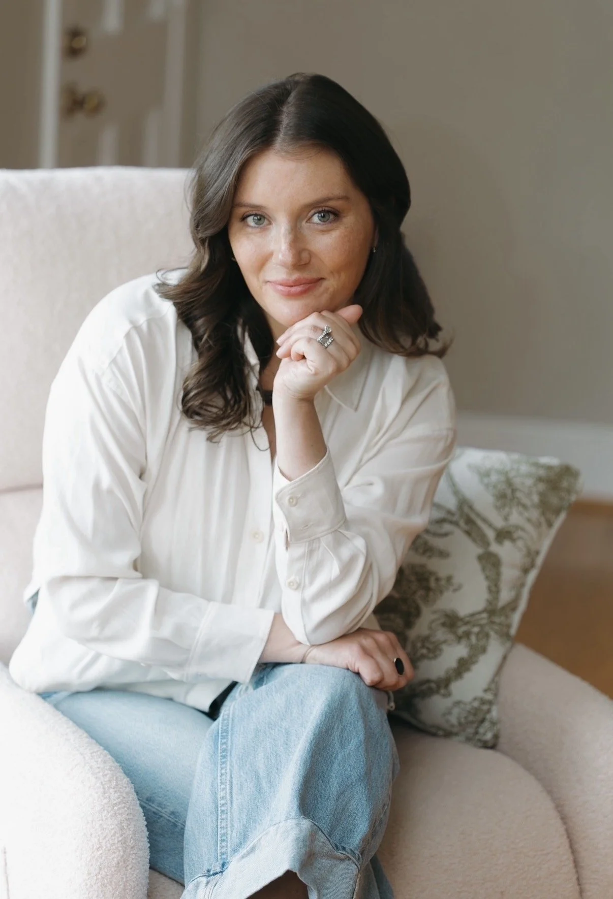 A woman with brown hair and blue eyes sitting on a light-colored sofa, resting her chin on her hand, wearing a white shirt and jeans, with a patterned pillow in the background.