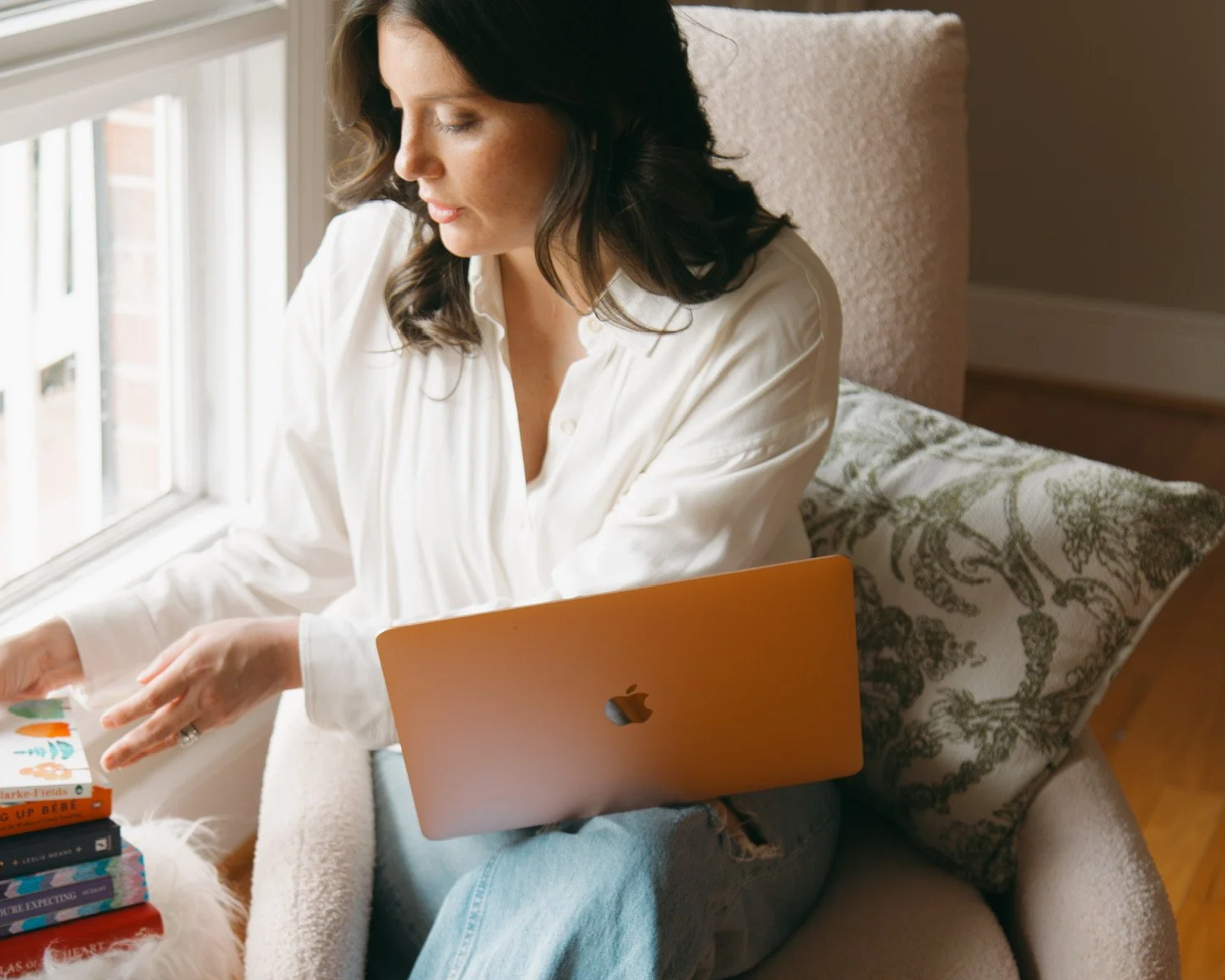A woman sitting on a beige armchair by a window with natural light, flipping through a children's book, with a closed Apple MacBook on her lap and a stack of books on her right.