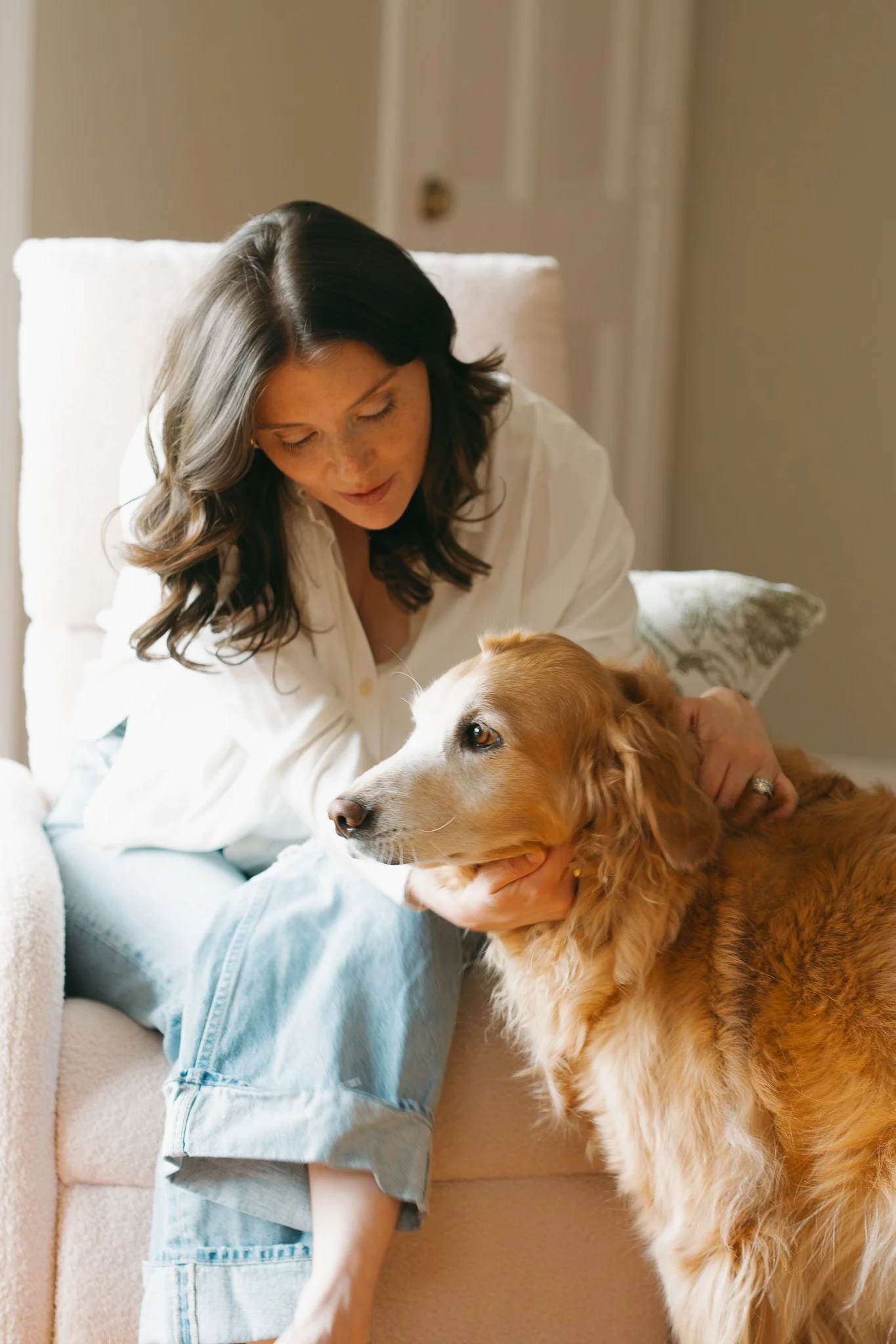 A woman with dark hair and freckles sitting on a beige couch, petting a large golden retriever with a white chest, in a room with a closed door in the background.