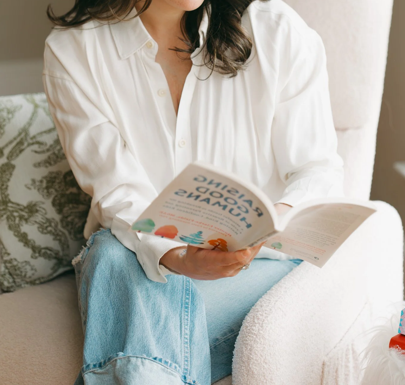 A woman with brown hair wearing a white button-up shirt and blue jeans is sitting on a cream-colored sofa with patterned pillows, reading a colorful book titled "A Good Time Playground."