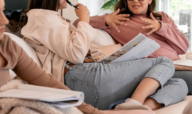 Two women sitting on a couch, engaged in a conversation, one holding papers and the other gesturing with her hands.