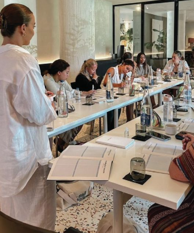 A woman stands at the front of a meeting room, with a large group of people seated at a long table, engaged in a discussion or presentation. The table has bottles of water, glasses, notebooks, and electronic devices.