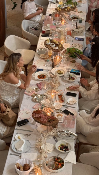 People dining and socializing at a long table decorated with candles, flowers, and food dishes.
