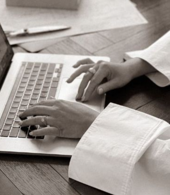 A person wearing a white shirt typing on a silver laptop on a wooden desk.
