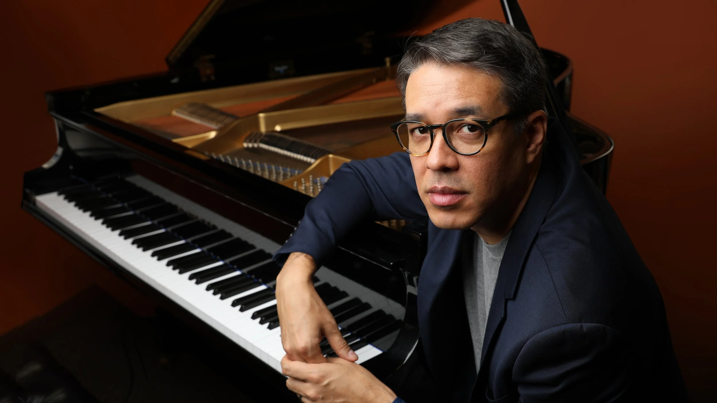 Composer, arranger, trombonist John Yao kneeling beside a grand piano