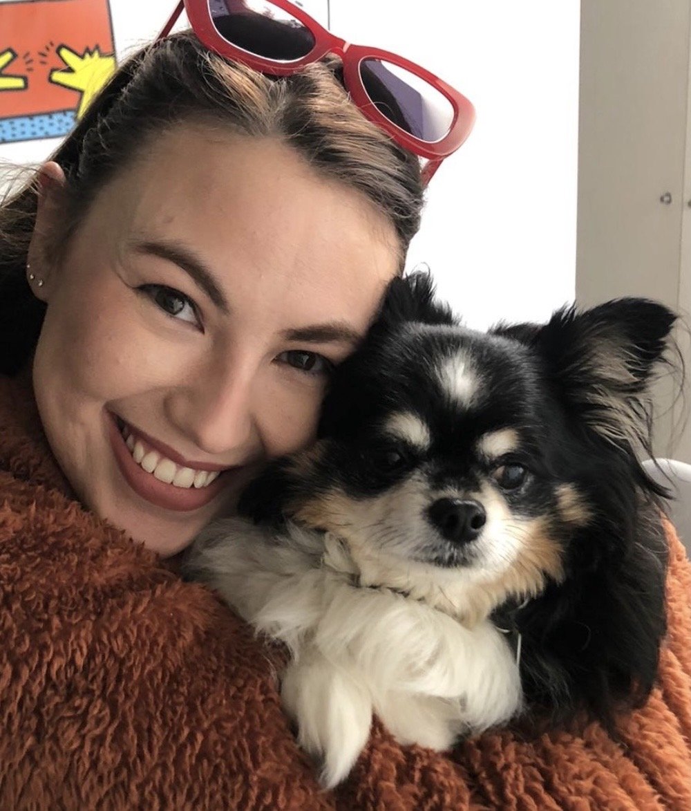 A smiling woman with red sunglasses on her head hugging a black and white dog with tan markings, both resting their heads together.
