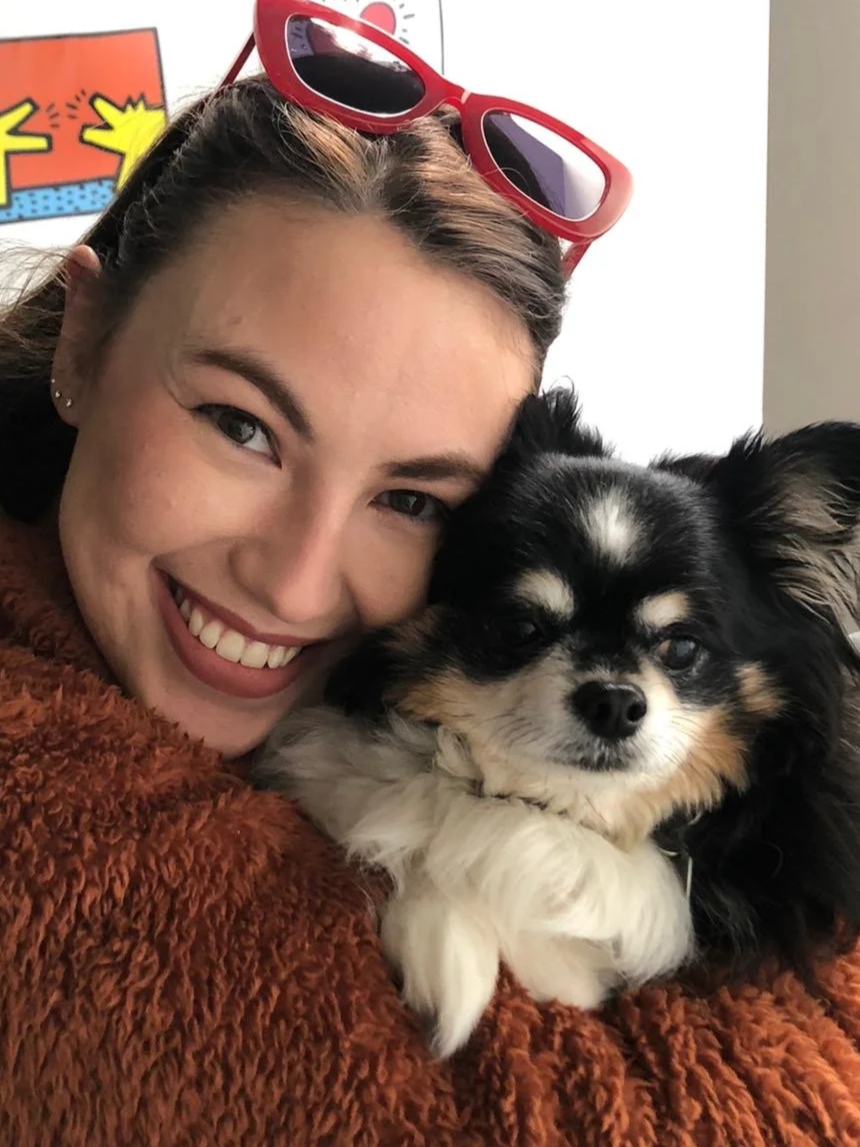 A smiling woman with red sunglasses on her head hugging a small black and white Chihuahua puppy.