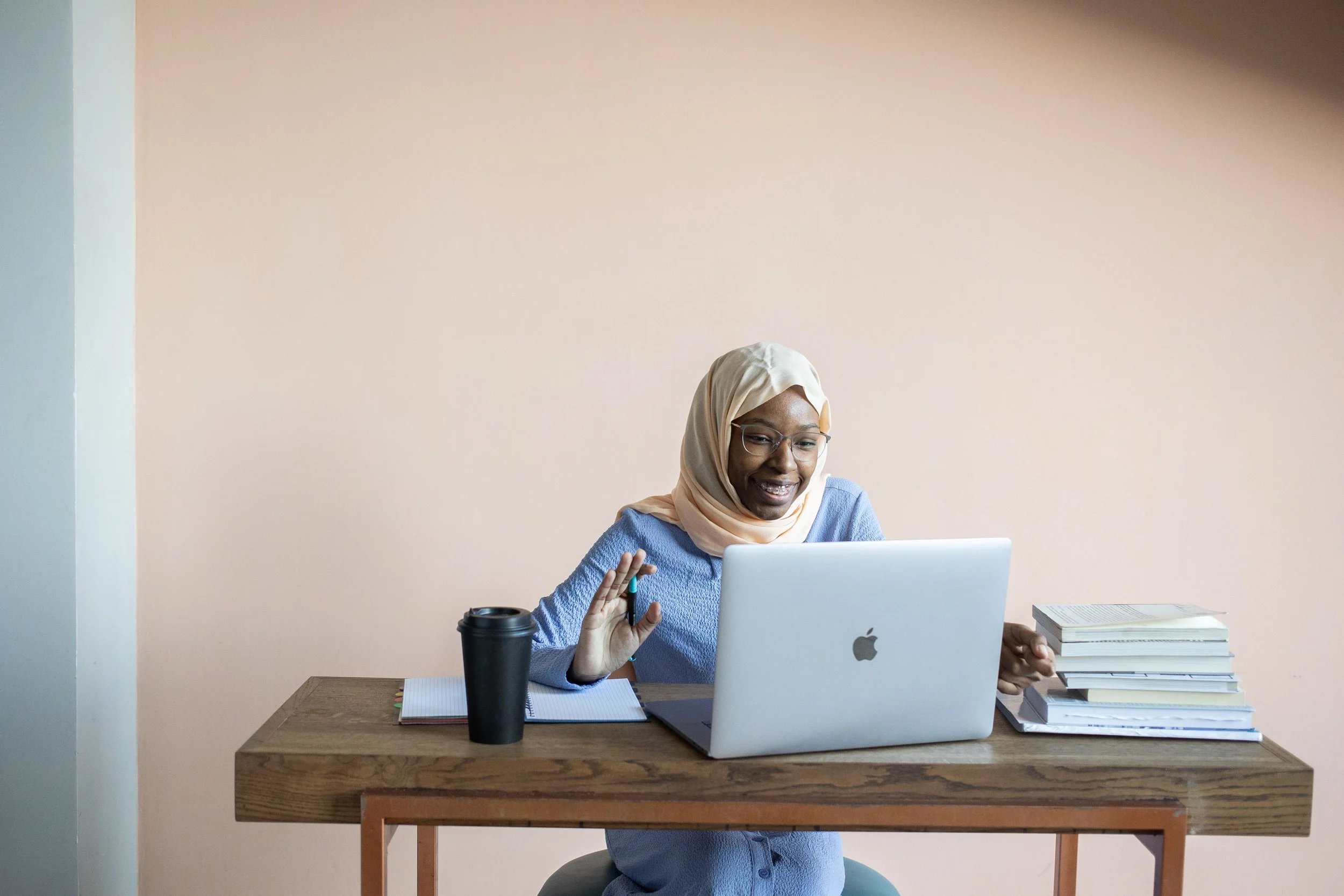 Woman wearing glasses and a hijab working on a laptop beside books and a notebook.