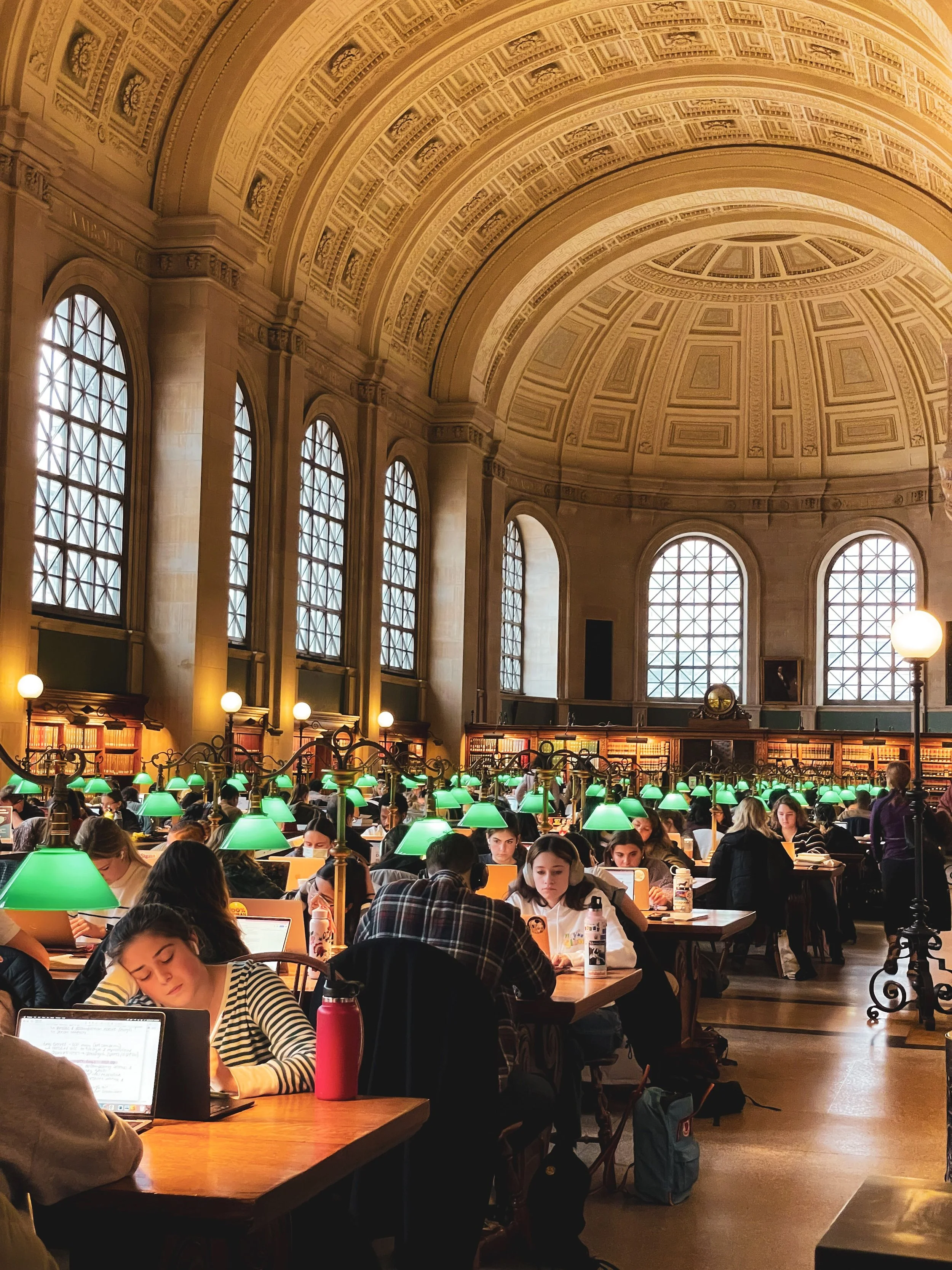 People reading and working on laptops at wooden tables in a grand library with arched windows, bookshelves, and green-shaded lamps.