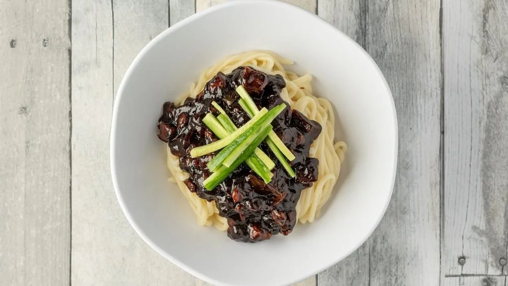 Bowl of Asian-style noodles topped with dark braised beef and garnished with sliced green cucumber on a wooden surface.