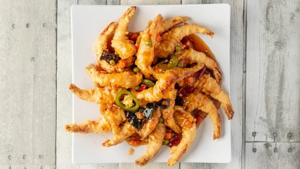 Plate of battered and fried shrimp topped with a spicy orange sauce and garnished with sliced jalapeños and chopped vegetables.