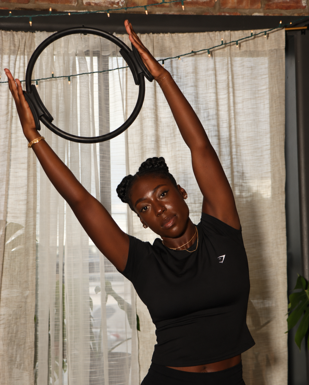 A woman with dark skin and braided hair in a bun stretches her arms above her head while holding a large black headphone circular headband in a room with beige curtains and string lights.