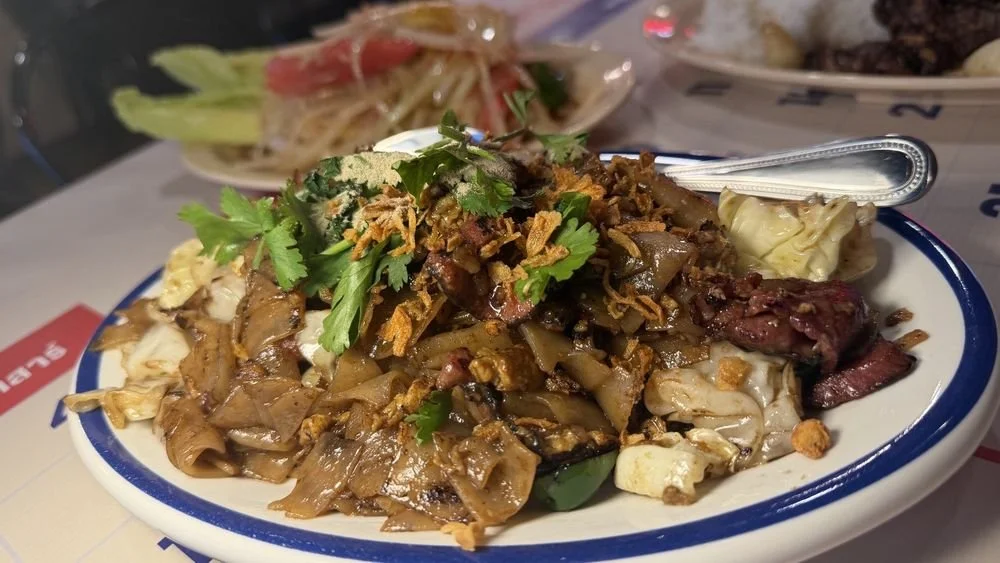 A plate of stir-fried noodles with beef, garnished with cilantro and fried shallots. In the background, there is a plate of salad with lettuce and tomato.