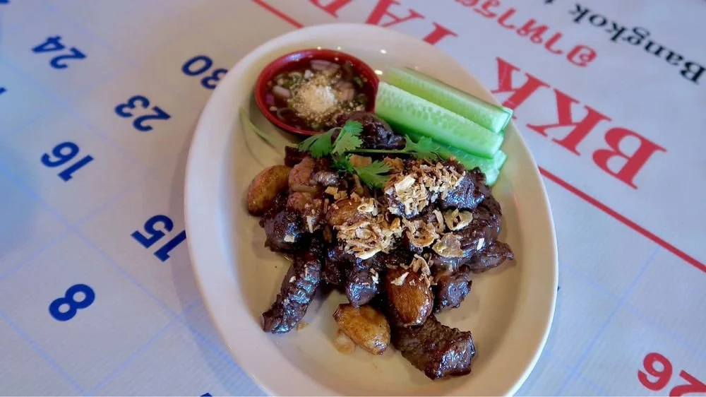 Plate of cooked beef or pork with garlic, served with cucumber slices, a cilantro garnish, and a small red bowl of dipping sauce on a table with printed text.