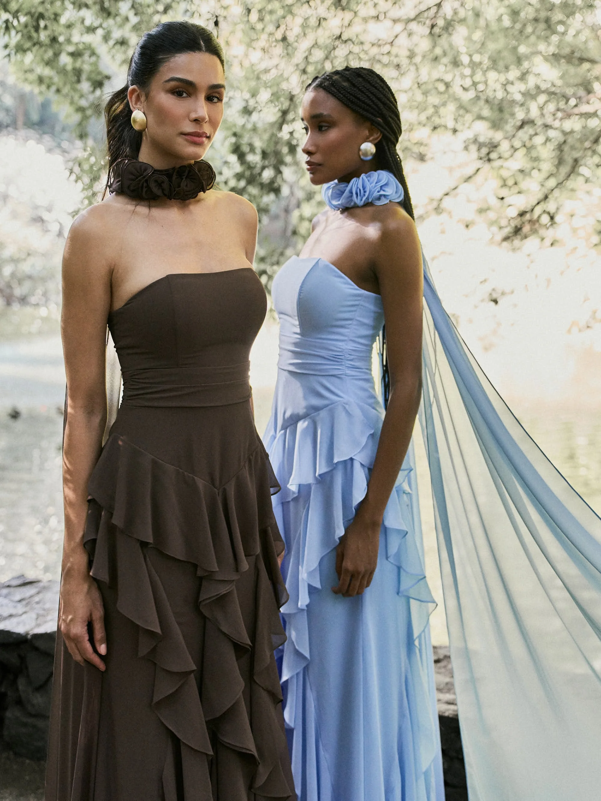 Duas mulheres com roupas elegantes e acessórios grandes em um ambiente natural ao ar livre, com árvores ao fundo, uma de costas, usando vestido azul com capa de tule e a outra usando vestido marrom escuro com babados.