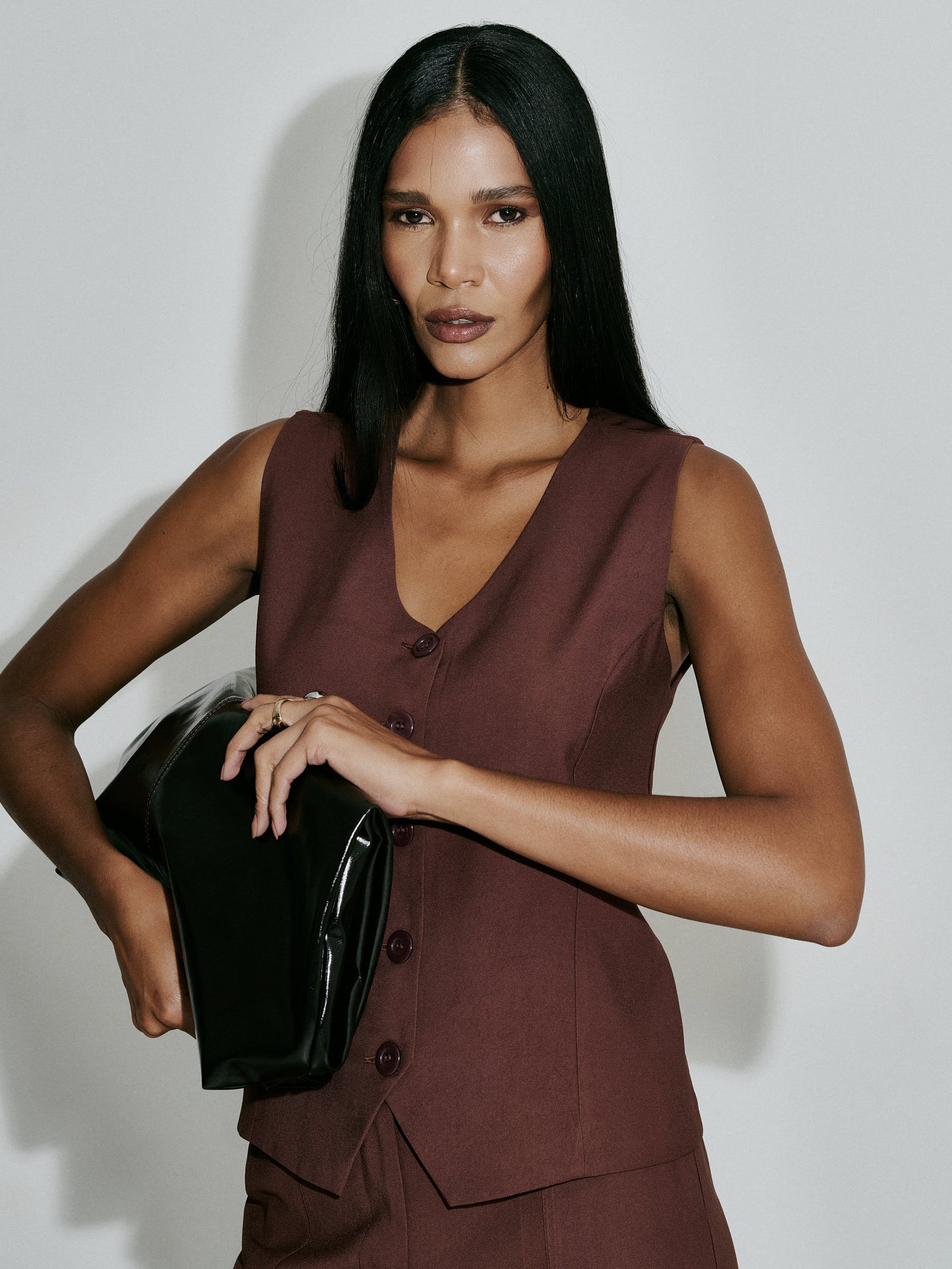Mulher com pele morena, cabelo liso e preto, vestindo vestido marrom com botões na frente, segurando uma bolsa preta, posicionada contra parede branca.