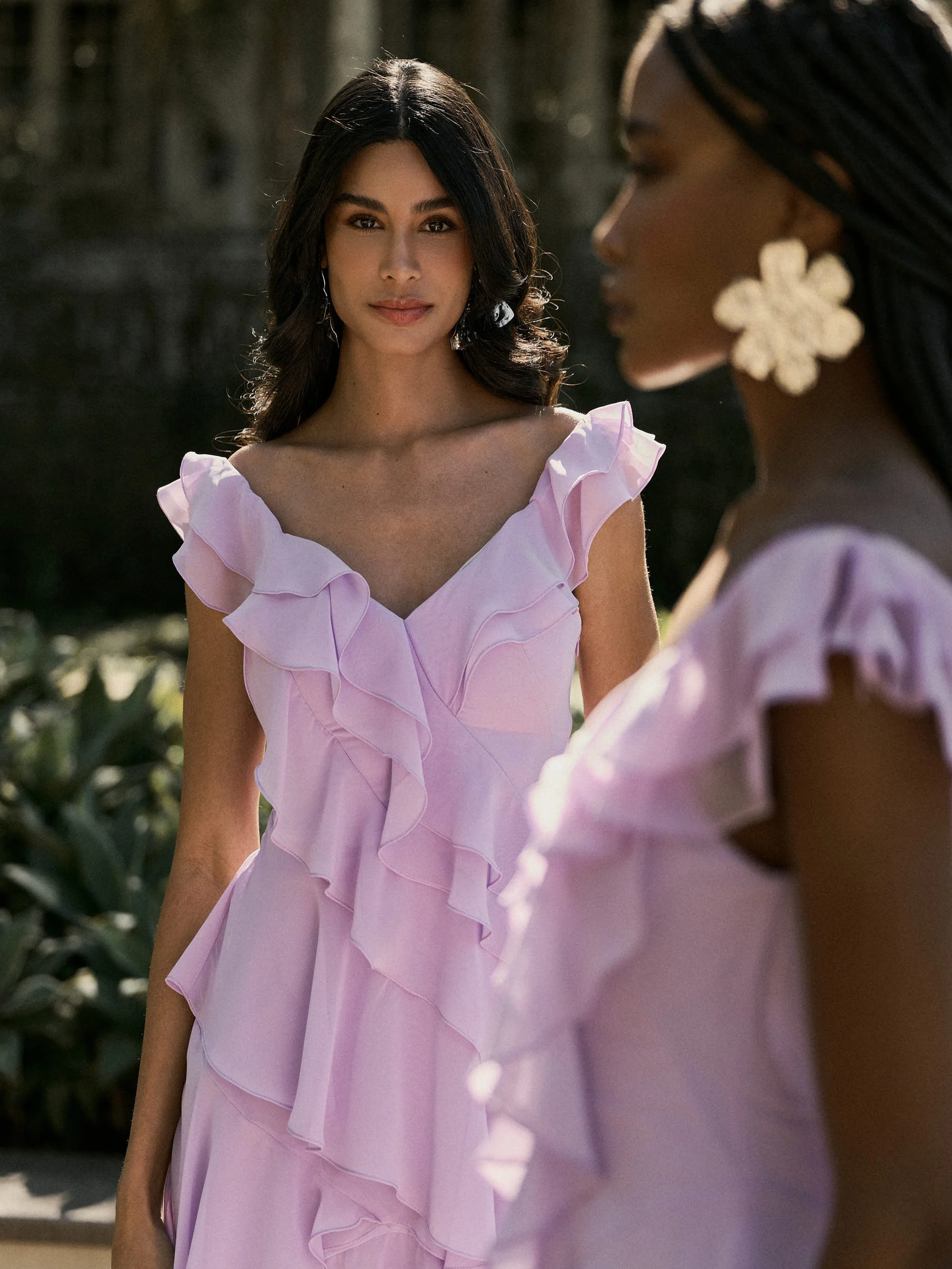 Duas mulheres usando vestidos rosas com detalhes de babados, uma delas com cabelo preto ondulado e a outra com cabelo trançado e brincos grandes em formato de flor, ao ar livre com vegetação ao fundo.
