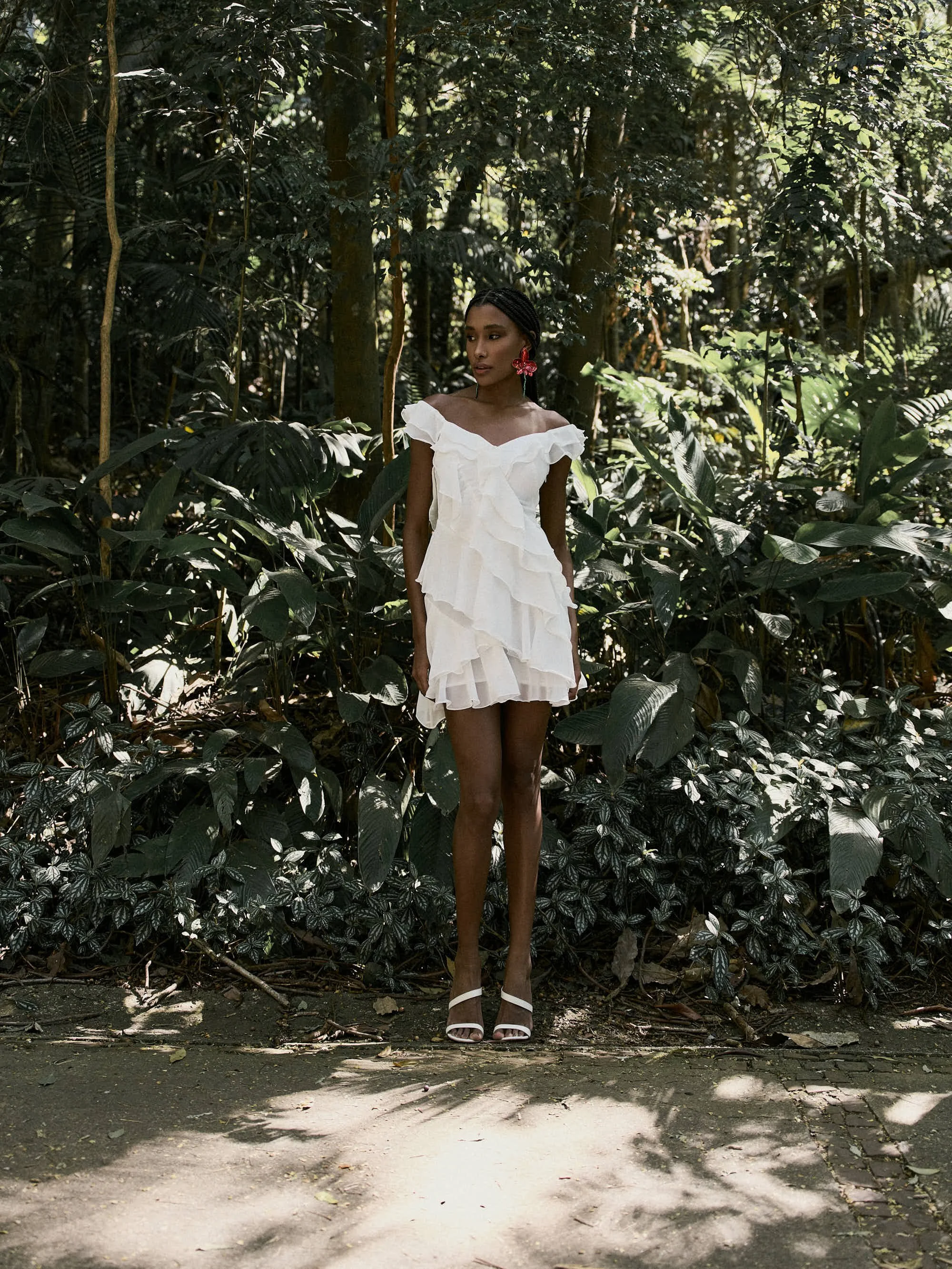 Mulher com pele escura vestindo vestido branco com babados, brincos vermelhos em forma de borboleta, sapatos brancos, de pé na calçada sob árvores e folhagem densa.
