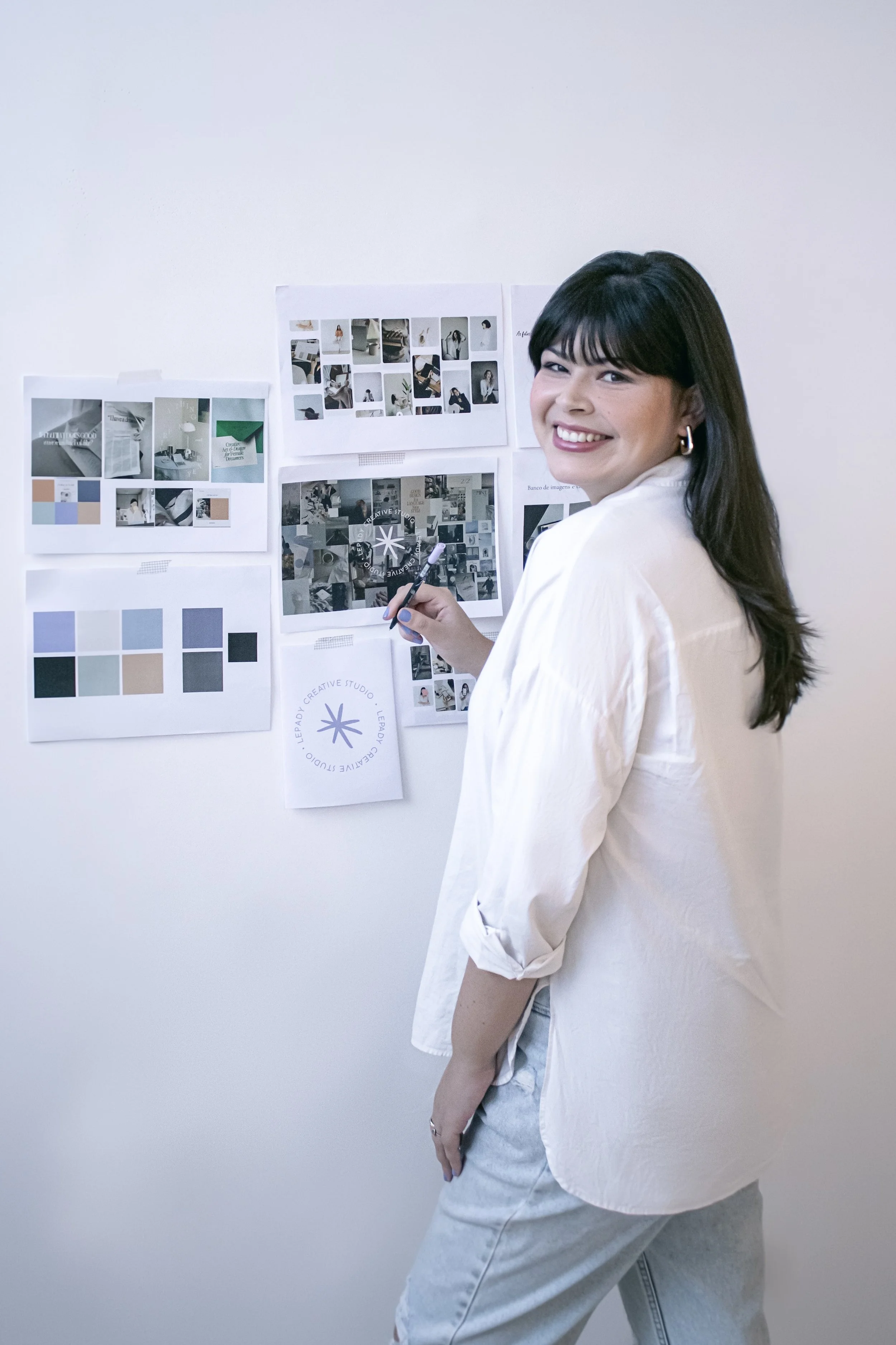 Mulher sorridente, de cabelos escuros e cacheados, usando uma blusa branca e jeans claro, posando ao lado de quadros com fotos e amostras de cores na parede branca.