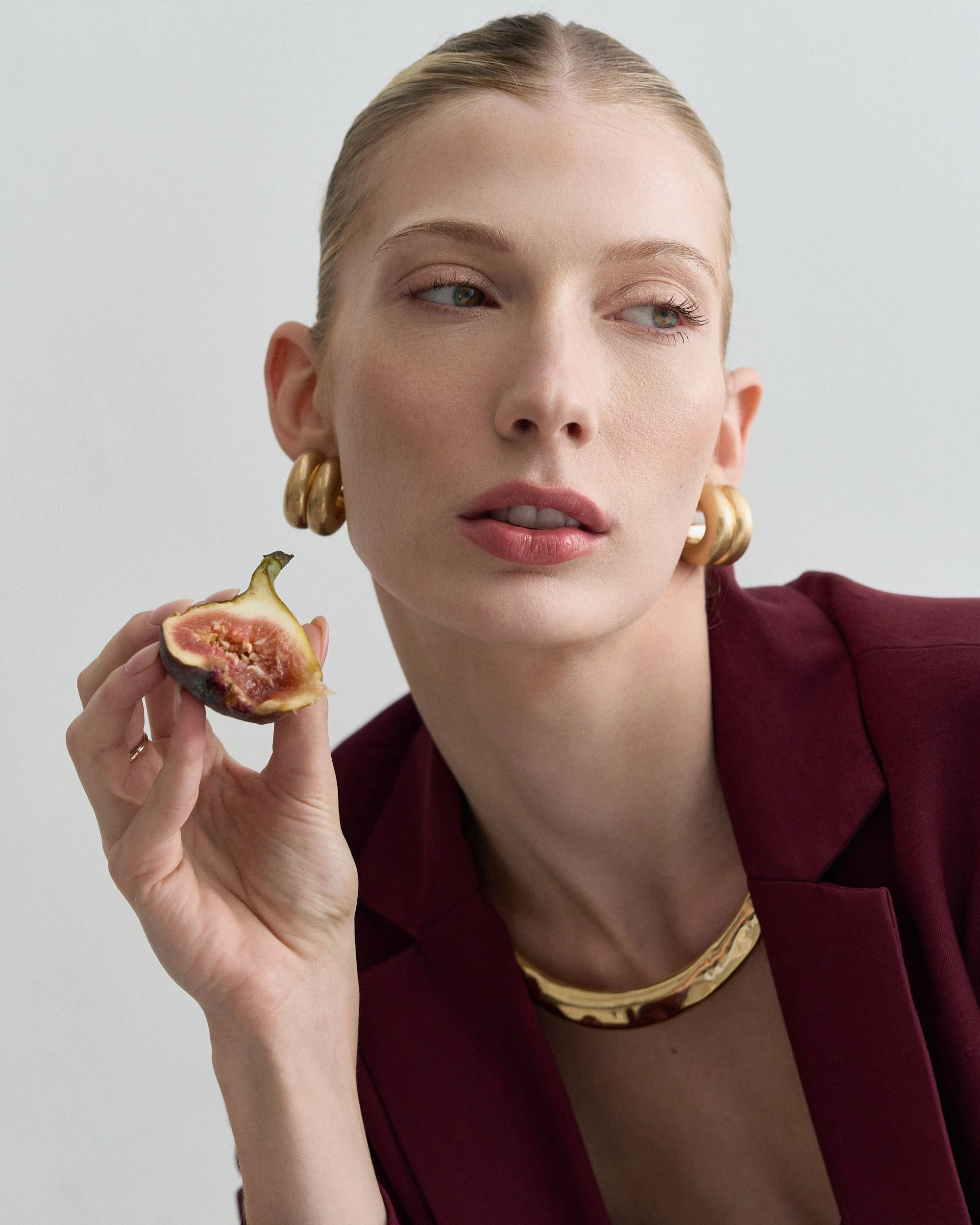 Mulher com cabelos presos, usando grandes brincos dourados e vestido vinho, segurando uma figo cortado, contra fundo branco.