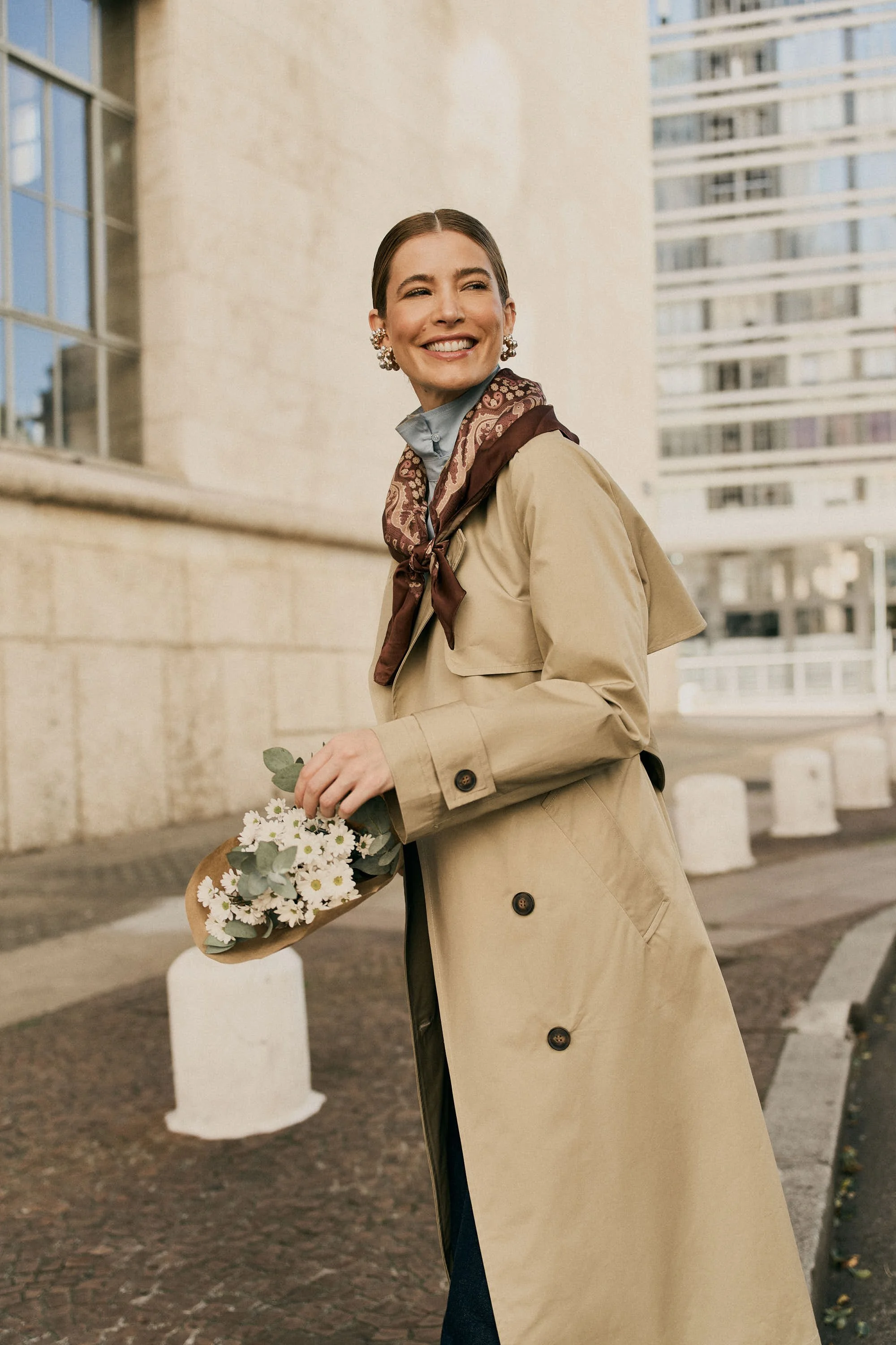 Mulher sorridente vestindo casaco bege, usando brincos grandes e lenço no pescoço, segurando buquê de flores brancas, ao ar livre em ambiente urbano.