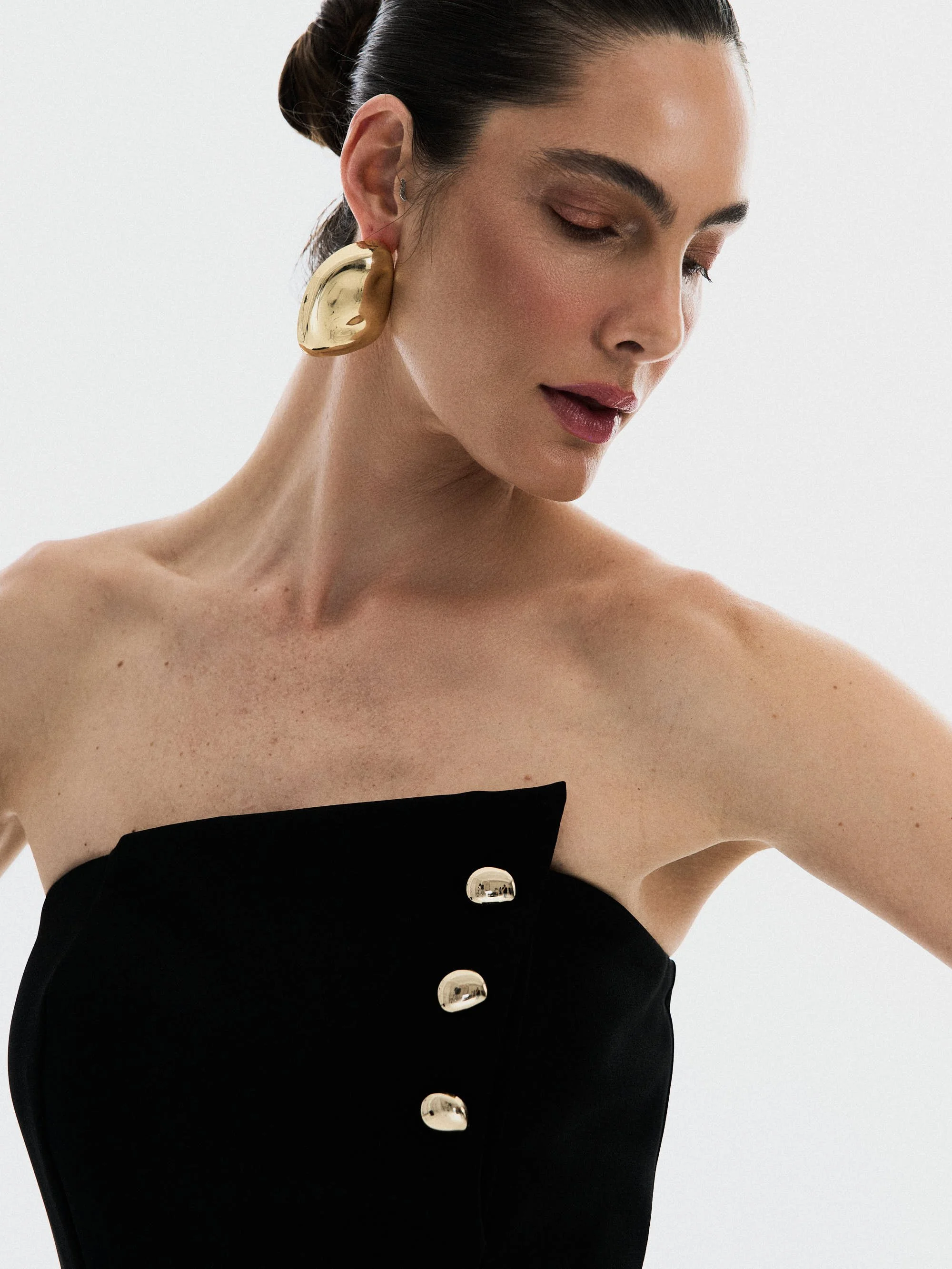 Mulher com cabelo preso, usando brincos grandes dourados e vestido preto com botões espelhados.