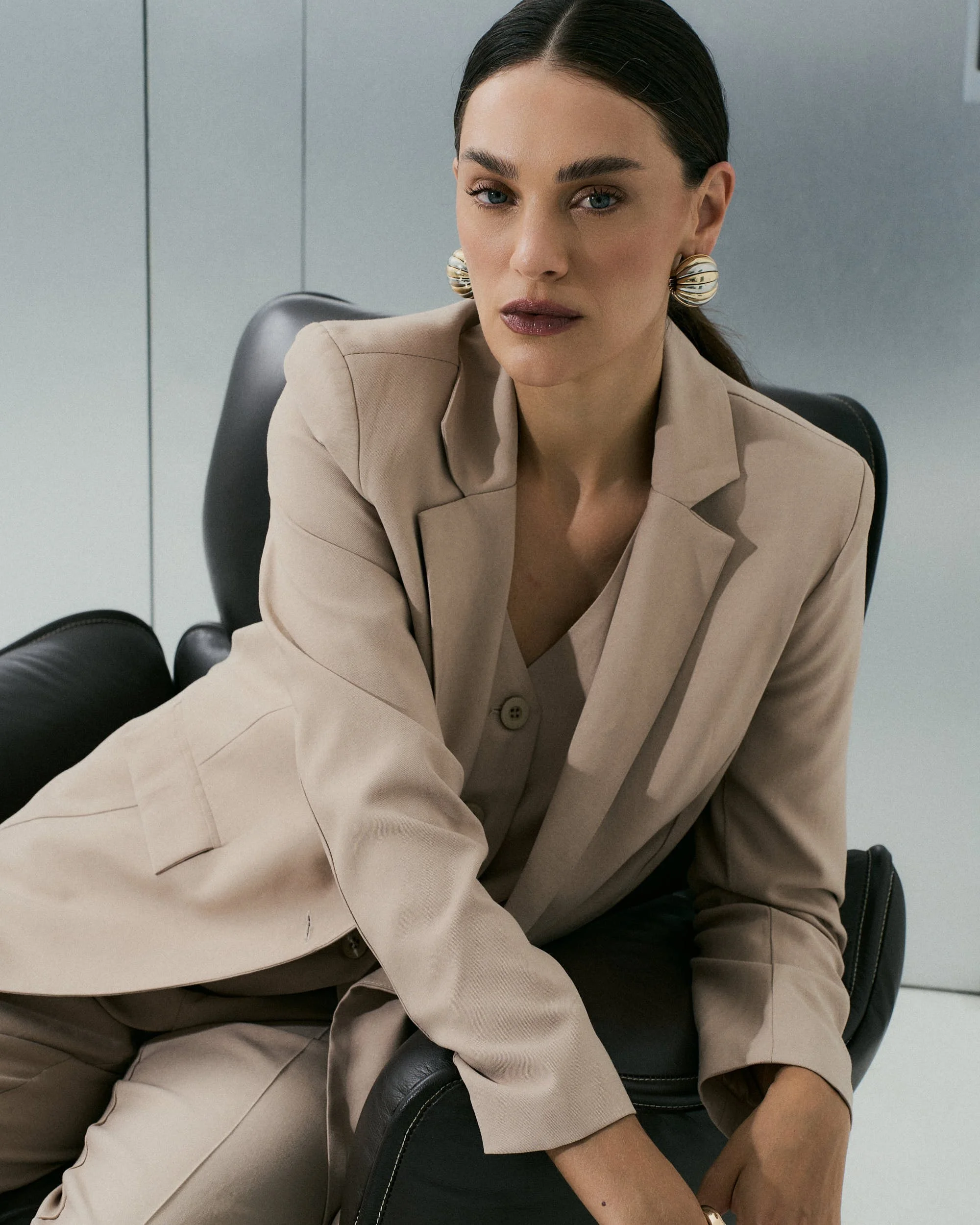Mulher com cabelo escuro, roupa bege, brincos grandes, sentada em cadeira de escritório, expressão séria