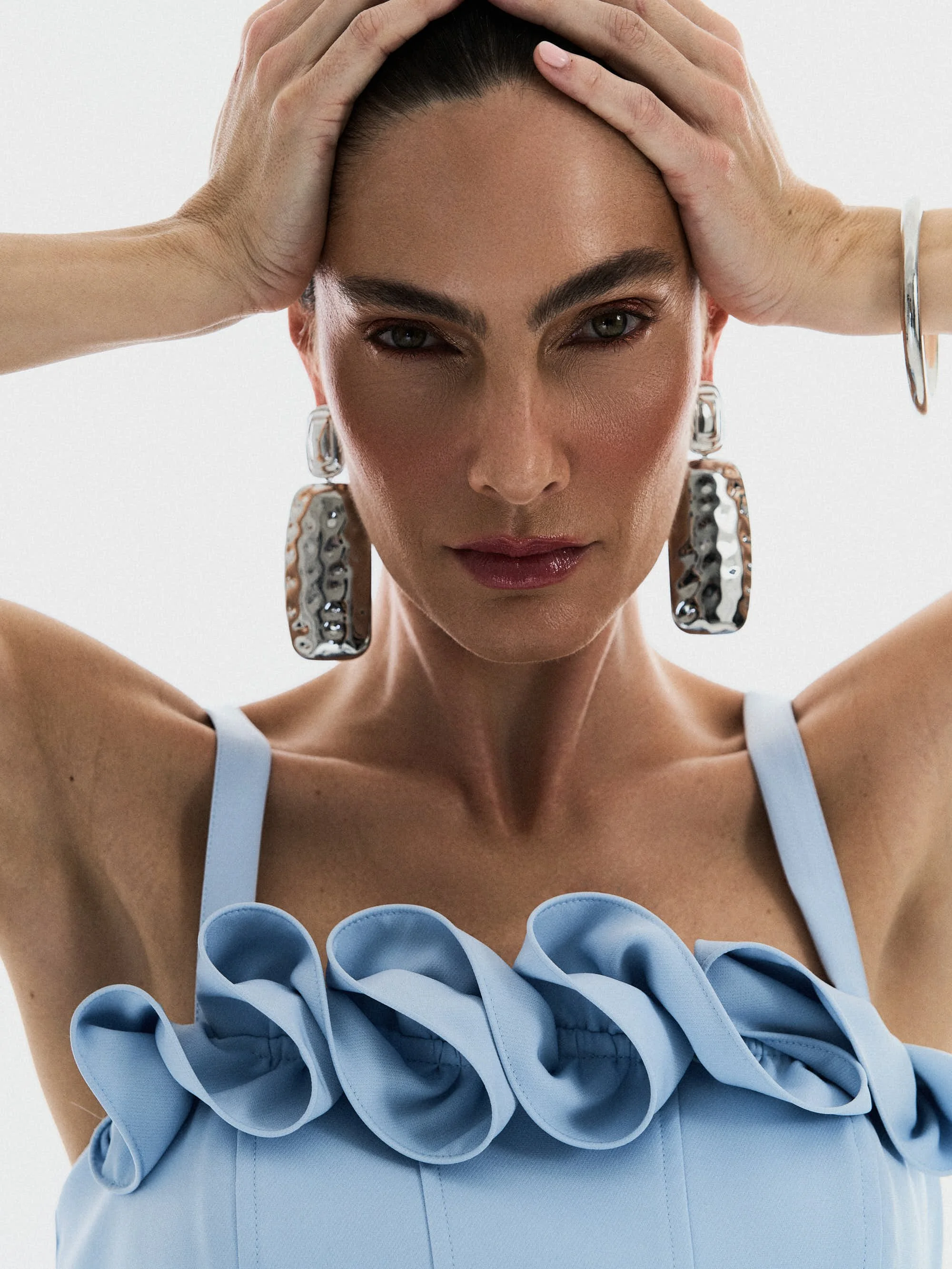 Mulher com brincos grandes de metal, usando vestido azul com detalhes dobrados, com mãos na cabeça e olhar direta para a câmera.