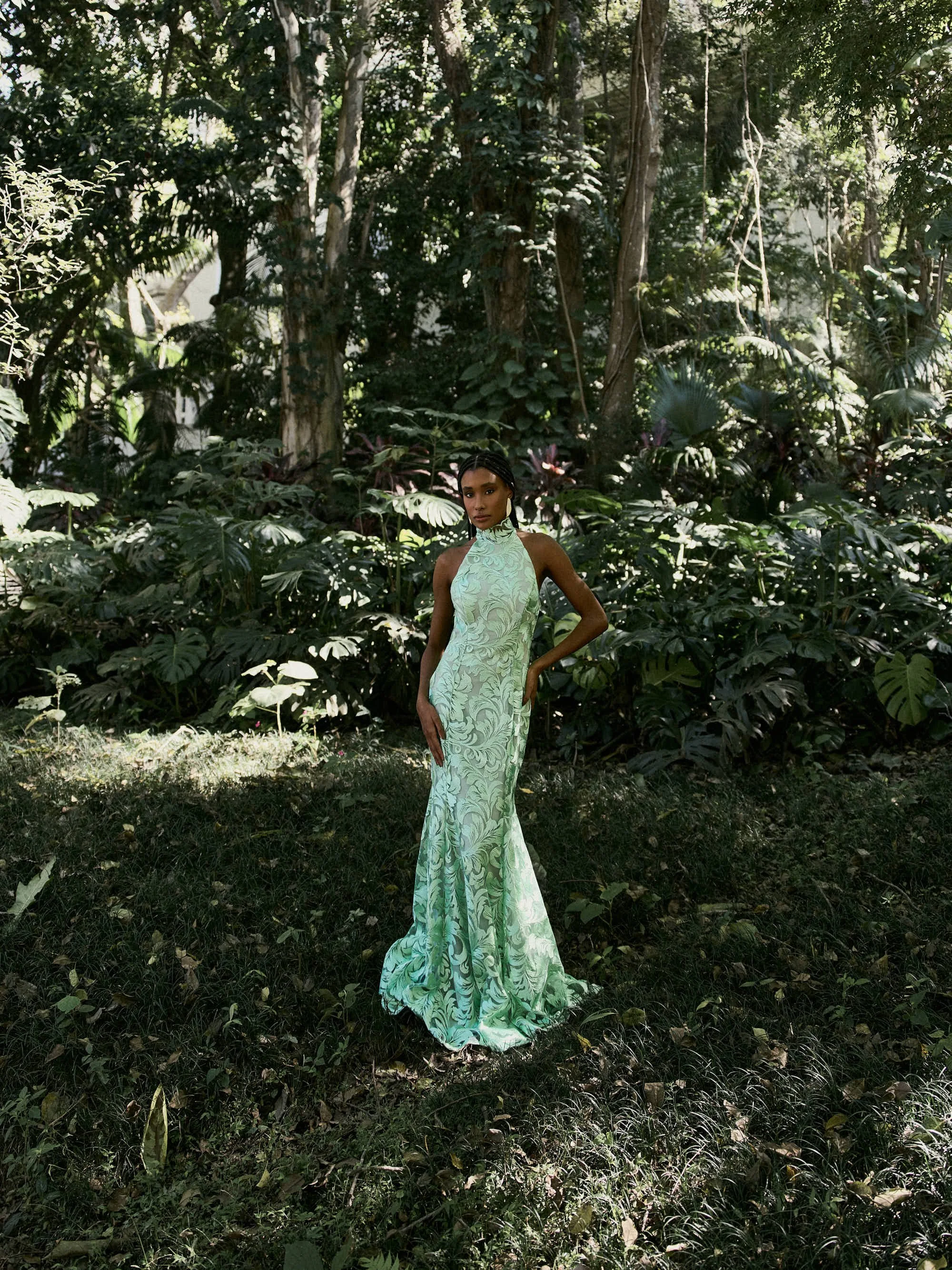 Mulher com vestido longo e de saia larga, de cor verde com estampa de folhas, posando em meio à vegetação densa de uma floresta