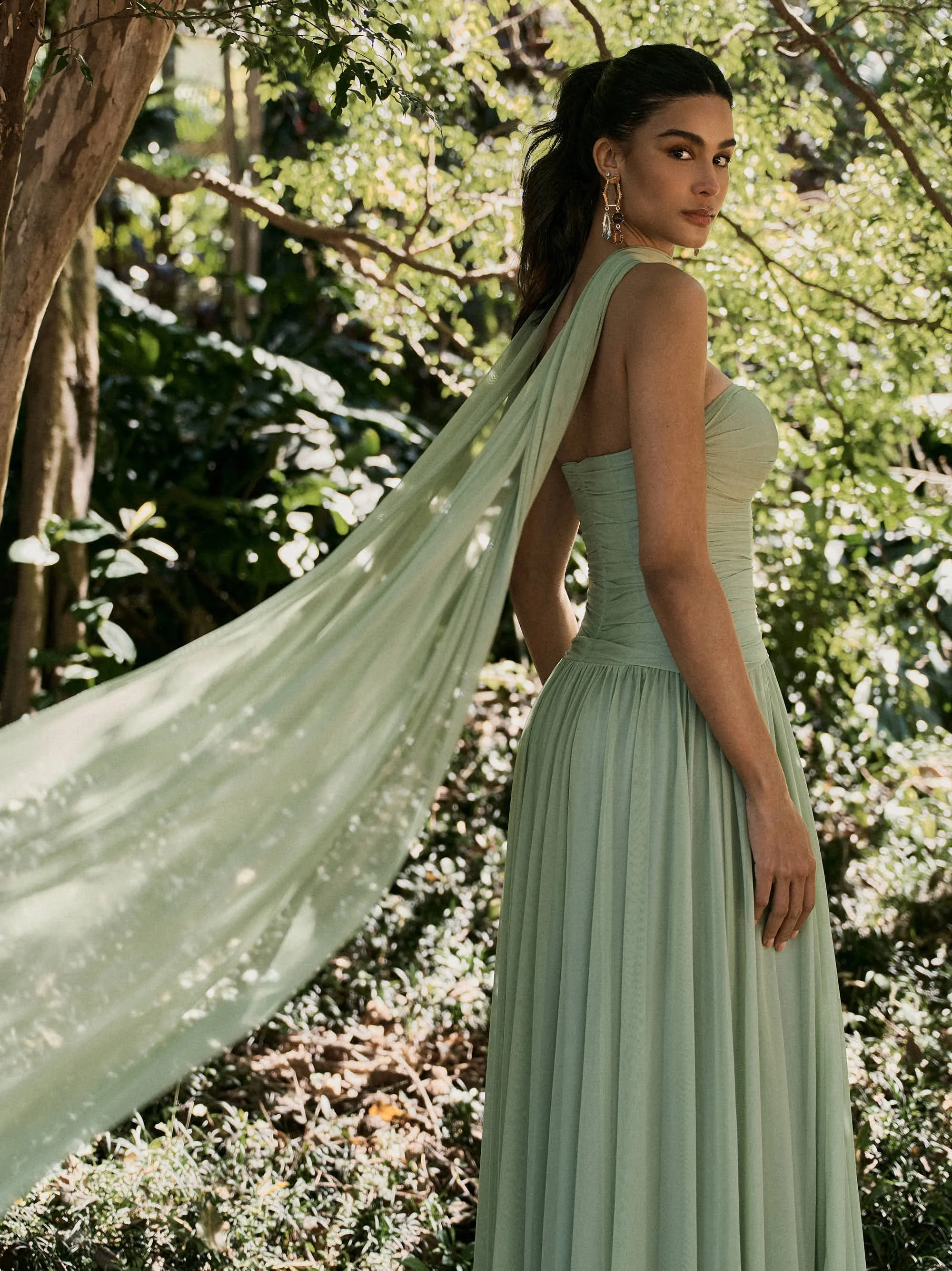 Mulher com vestido verde claro e brincos grandes numa área de floresta com árvores e folhagens ao fundo.